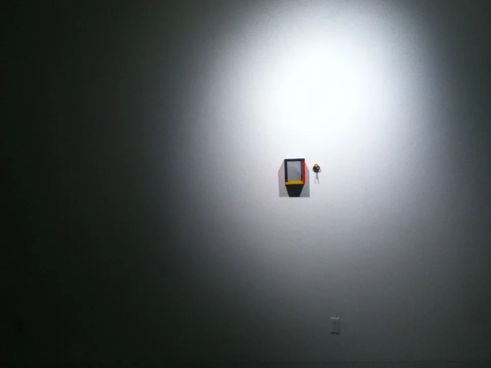 An art installation on a gray gallery wall featuring a small floating cabinet and a colorful flower bouquet hanging nearby.