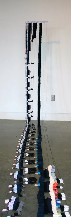 Art installation featuring a long, vertically hanging textile piece with various fabric strips and strings, creating a visual flow from top to bottom against a blank wall and floor.