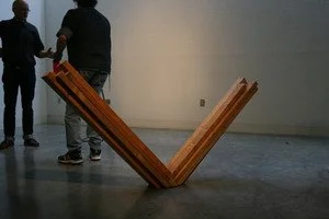 Two people standing near a large wooden V-shaped structure in a gallery or indoor space.