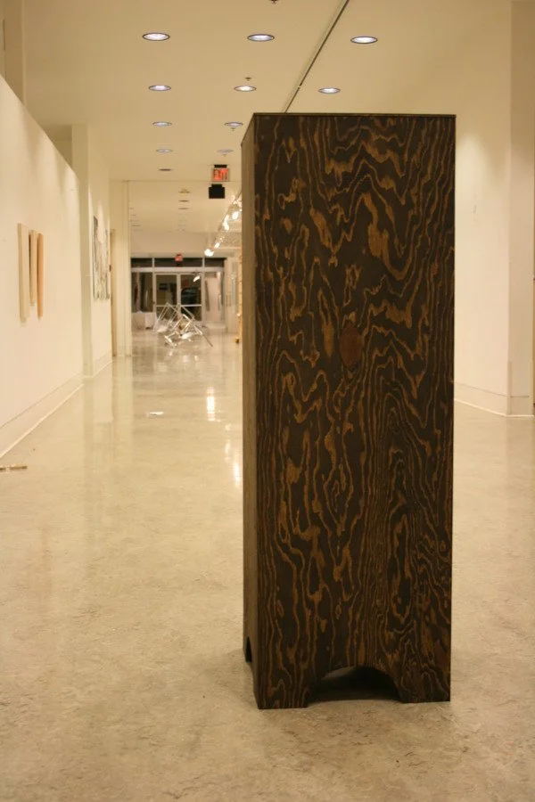 A dark, textured wooden sculpture or chair standing on terrazzo floor in a brightly lit art gallery or museum hallway with white walls and artwork.