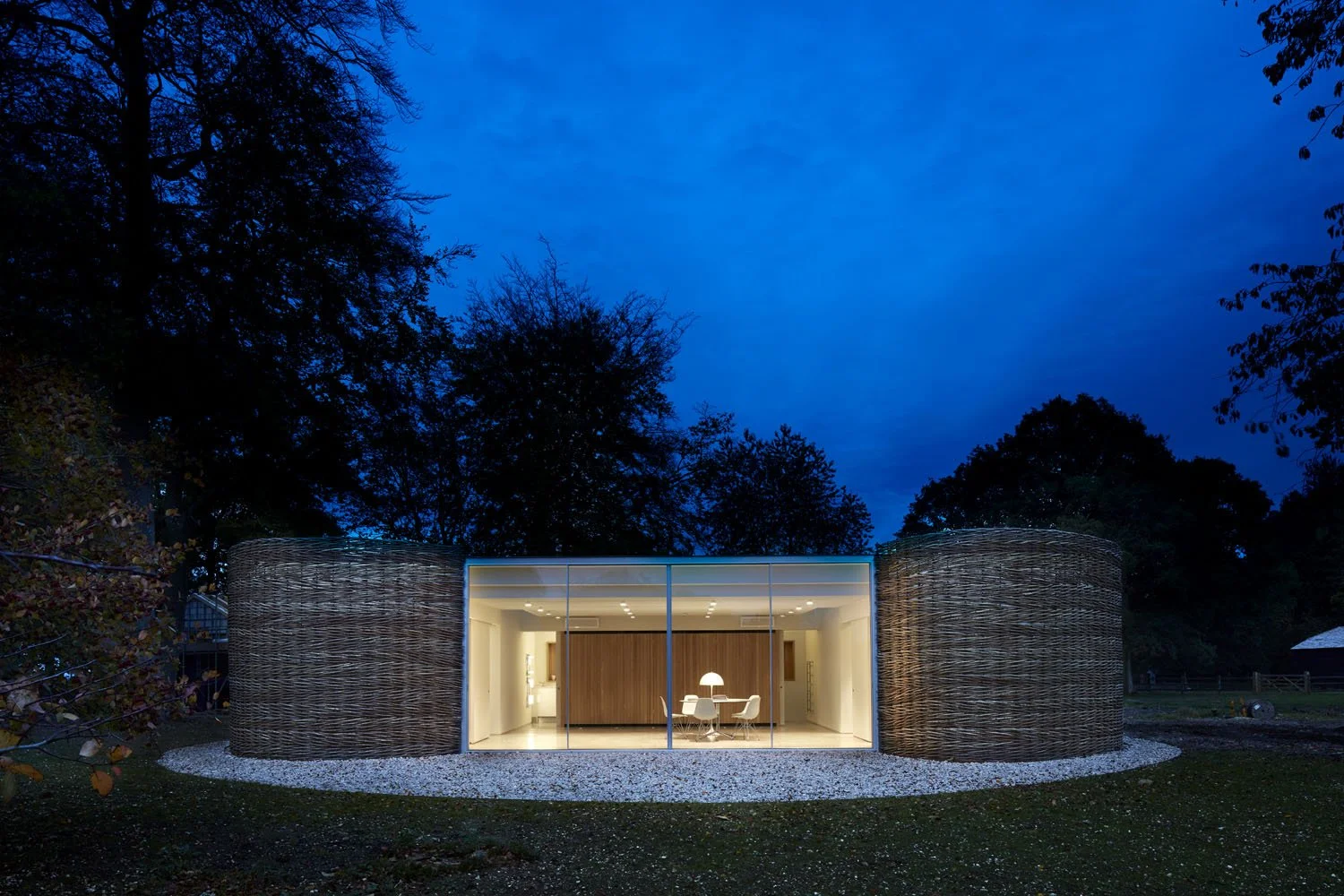 Modern house with glass front and woven exterior walls, illuminated inside, surrounded by trees at dusk.