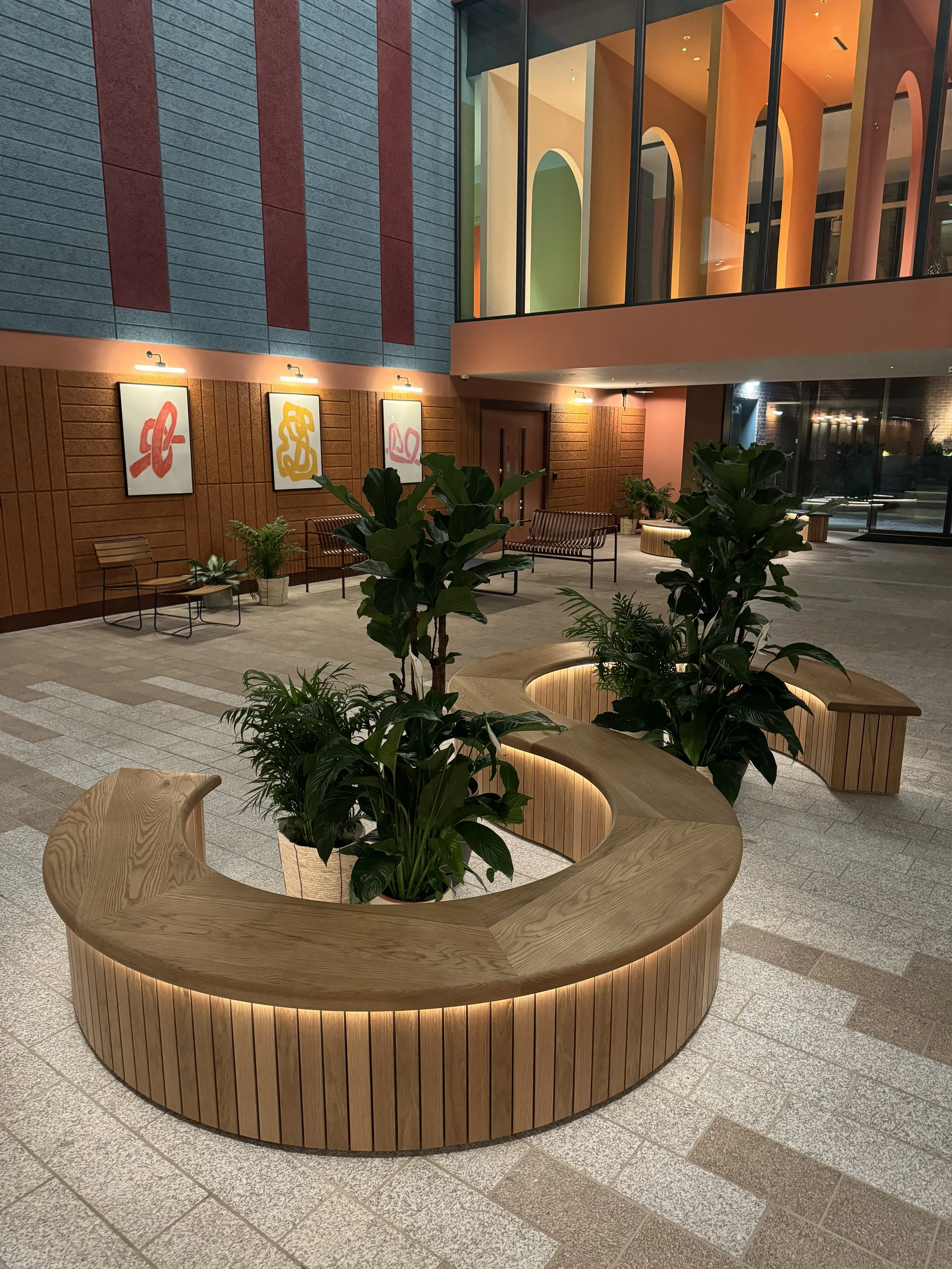 Modern indoor lobby with wooden curved seating and potted plants, colorful abstract artwork on the wall, and large windows with vibrant geometric shapes outside.