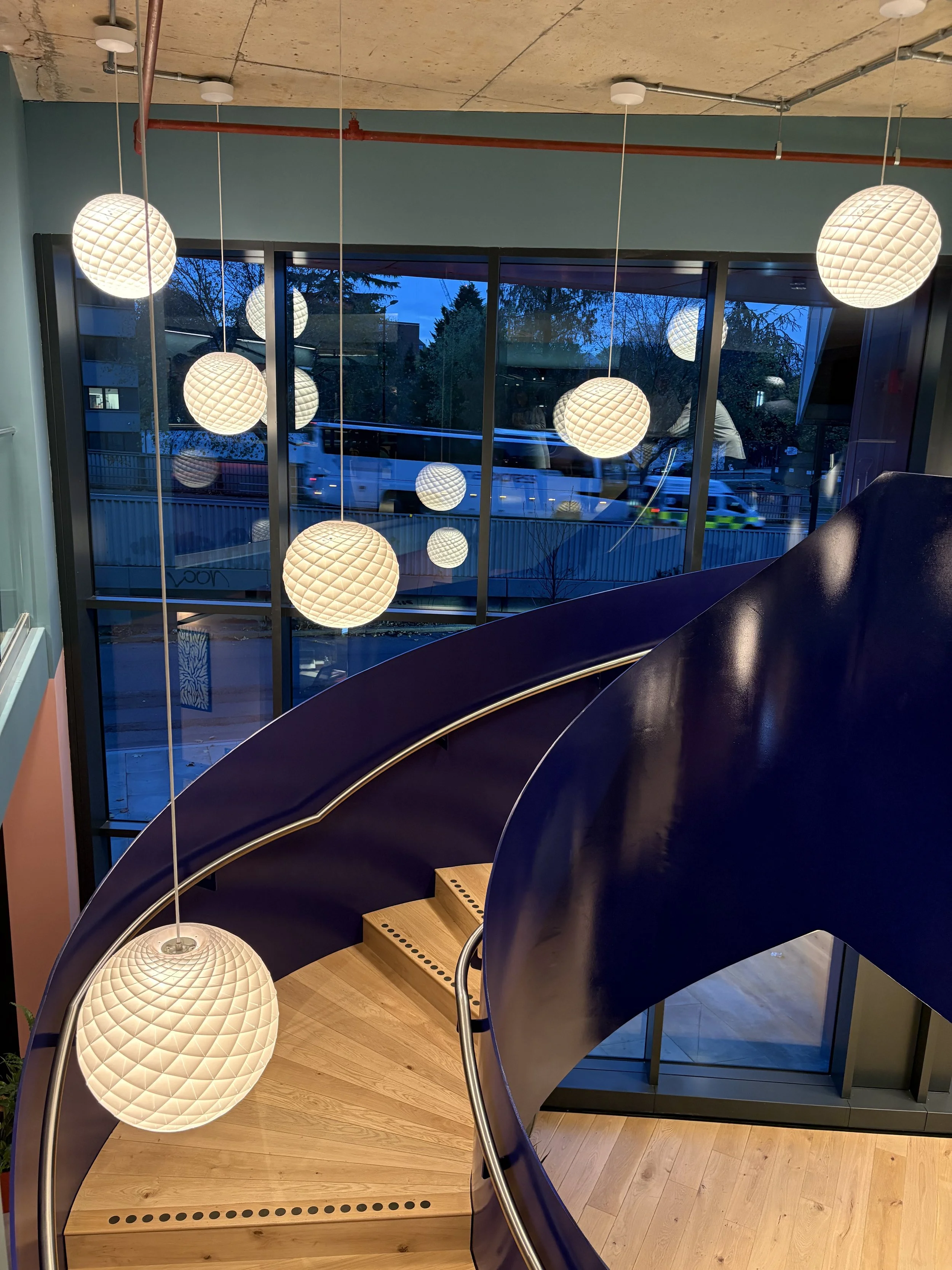 Interior view of a staircase with wooden steps, black railing, modern hanging pendant lights, and large glass windows showing trees and vehicles outside during evening.