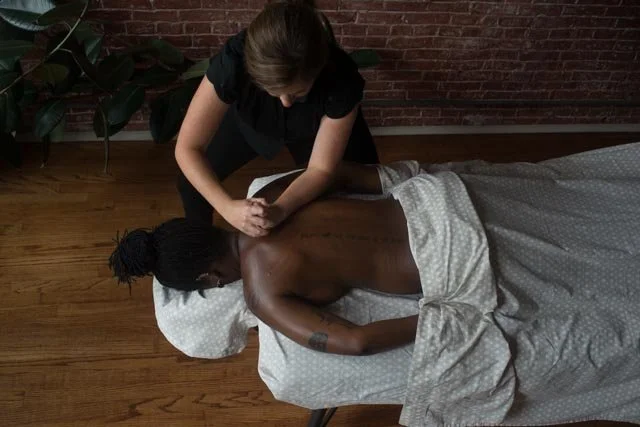 A person with dark skin lies face-down and relaxes as a massage therapist applies pressure to their back