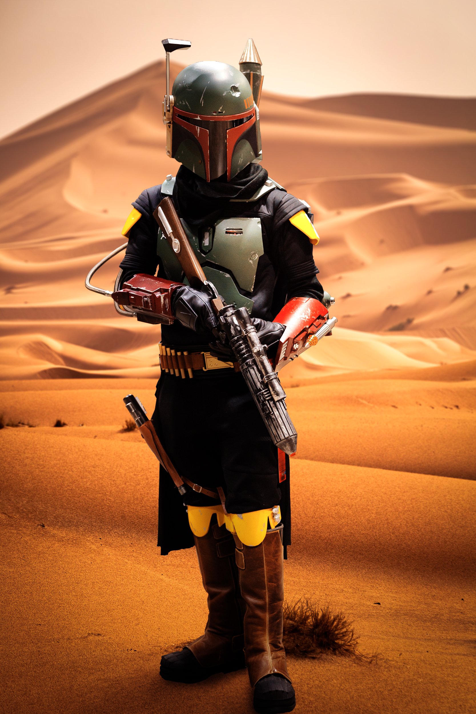 The image is a full body cosplay portrait of a man dressed as a Boba Fett from Star Wars. He stands in a battle ready pose, embodying the spirit of the renowned bounty hunter. The background depicts a desert landscape with vast sandy dunes and toweri