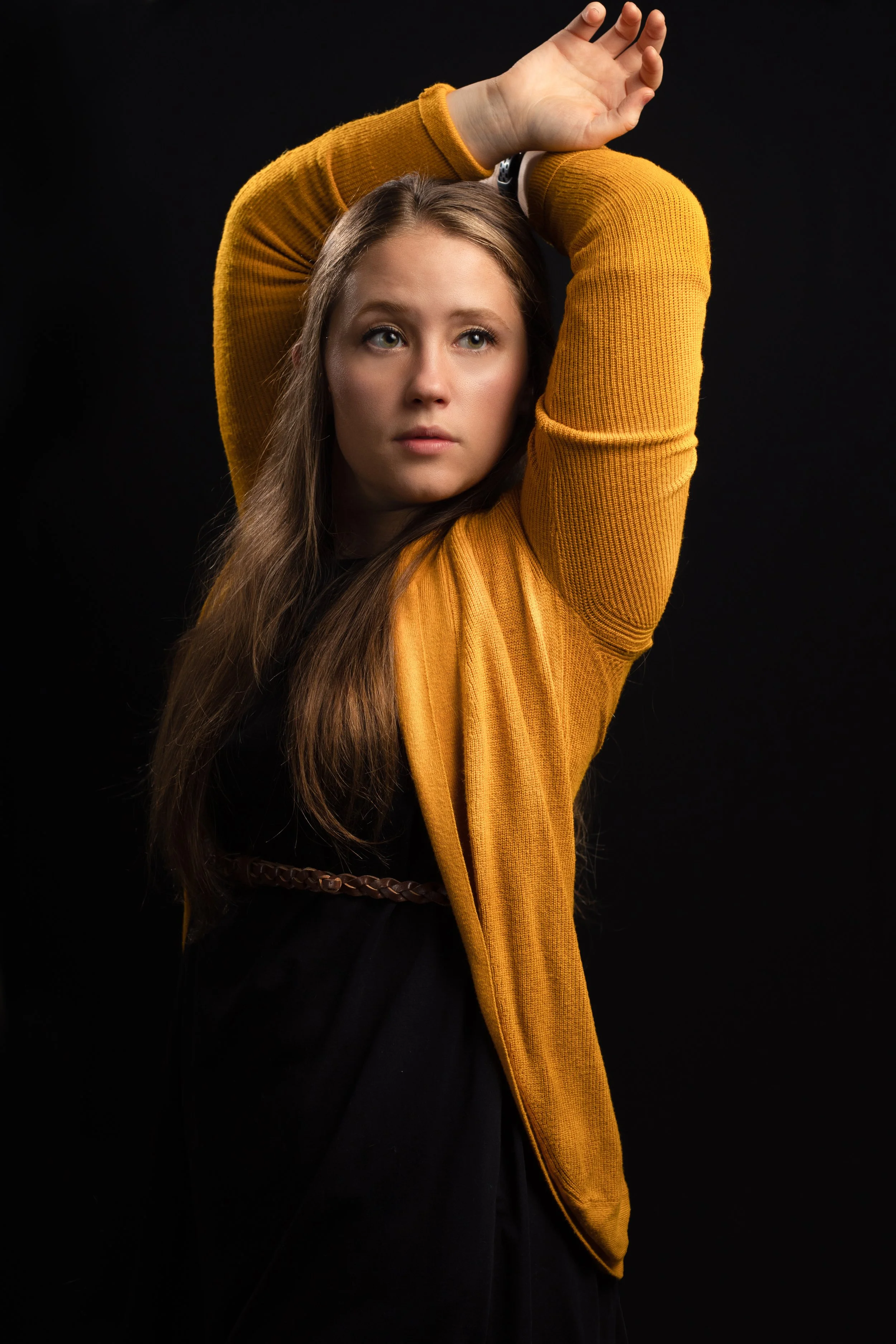 A white woman wearing a yellow cardigan strikes a fashion pose with her arms raised above her head. She gazes off-camera, exuding a sense of confidence and style.