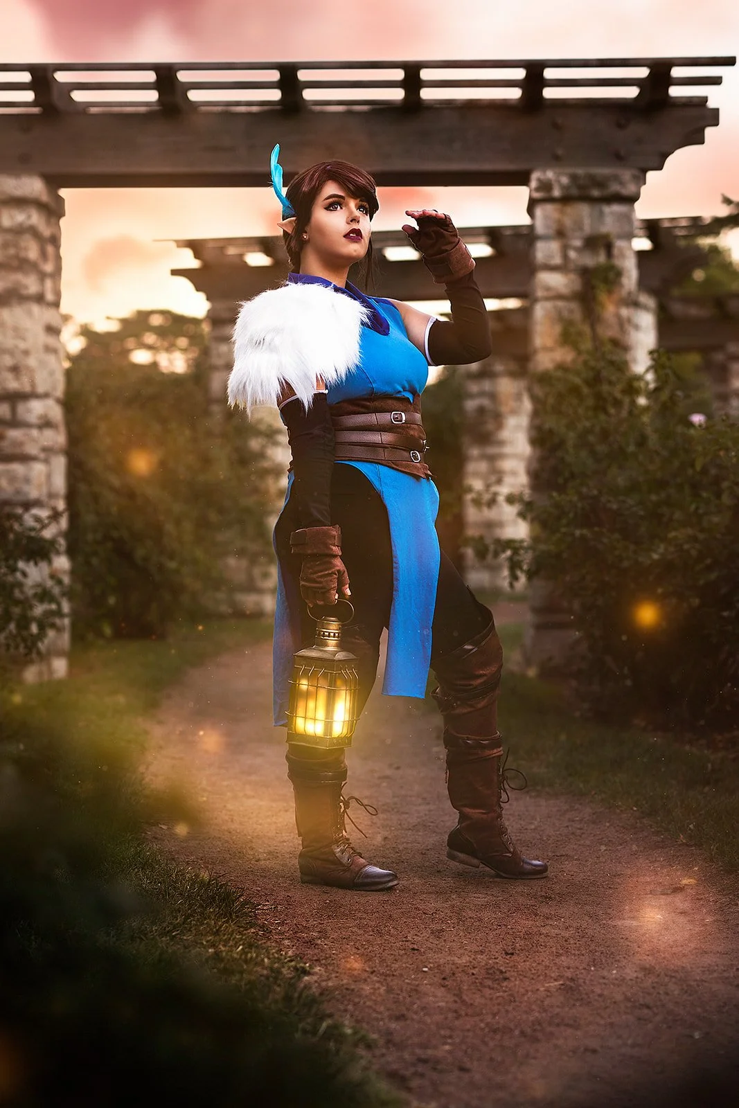 The photo features a woman in a cosplay costume, embodying the character Vex'Ahlia from Critical Role. She strikes a pose with a fashion-forward flair, exuding confidence. In her hand, she holds a lantern emanating sparkling light. Her gaze is direct
