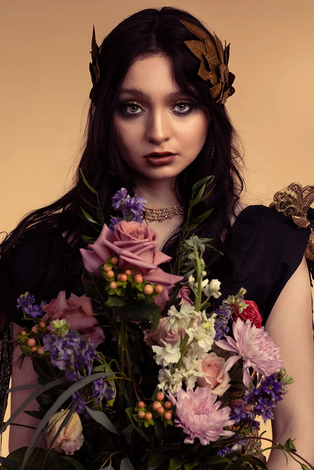 A captivating beauty portrait of a woman with black hair and a black dress. She adorns a golden Greek crown, adding an element of regal elegance to her appearance. Her gaze is soft and at the camera. She holds a bouquet of flowers, which adds a delic