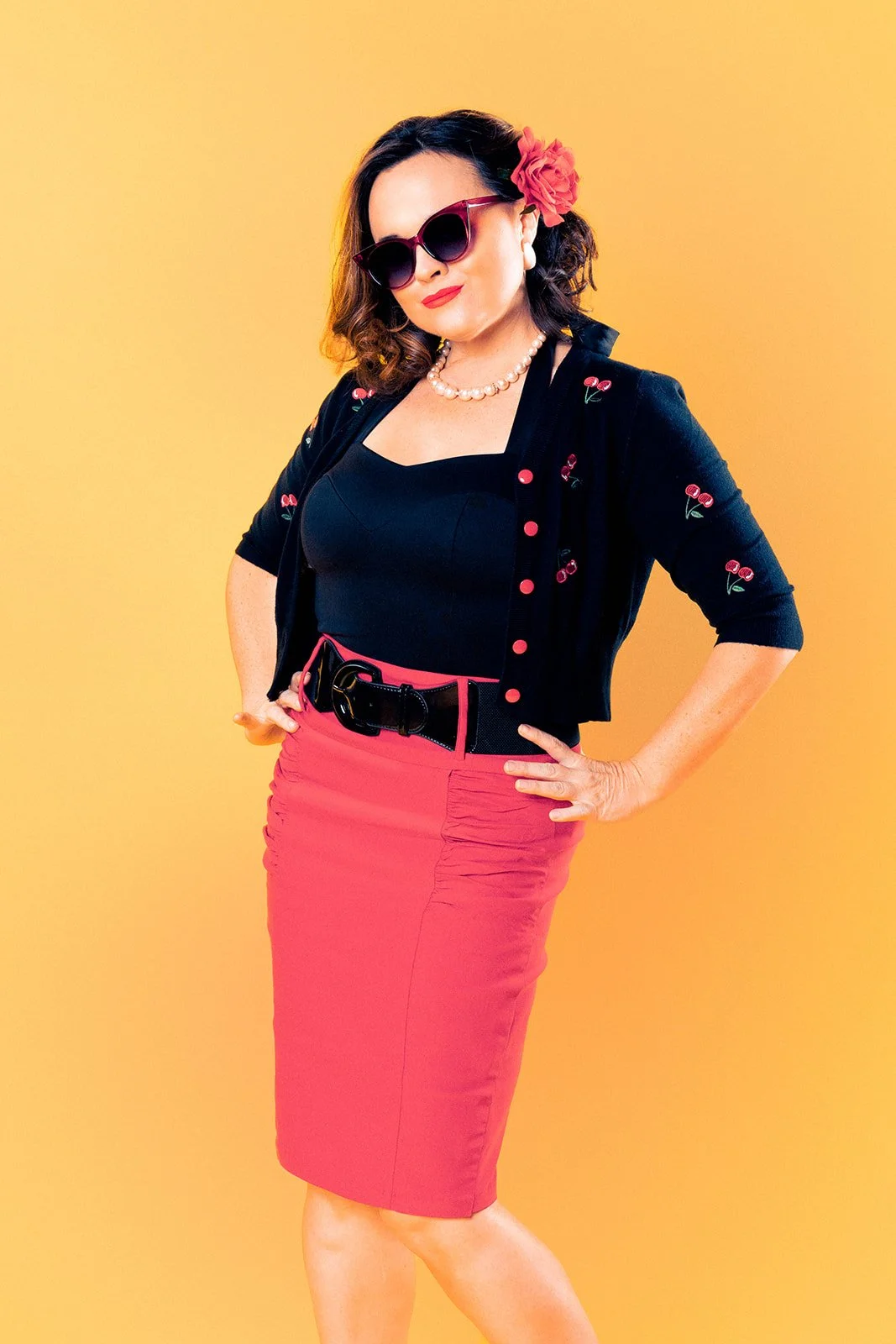 A full body studio portrait of a white woman in a 1950's pinup style, posing in a classic pinup pose with her hands on her hips.  She embodies the classic pinup aesthetic, with retro fashion and styling. The background has a soft yellow tone, adding 