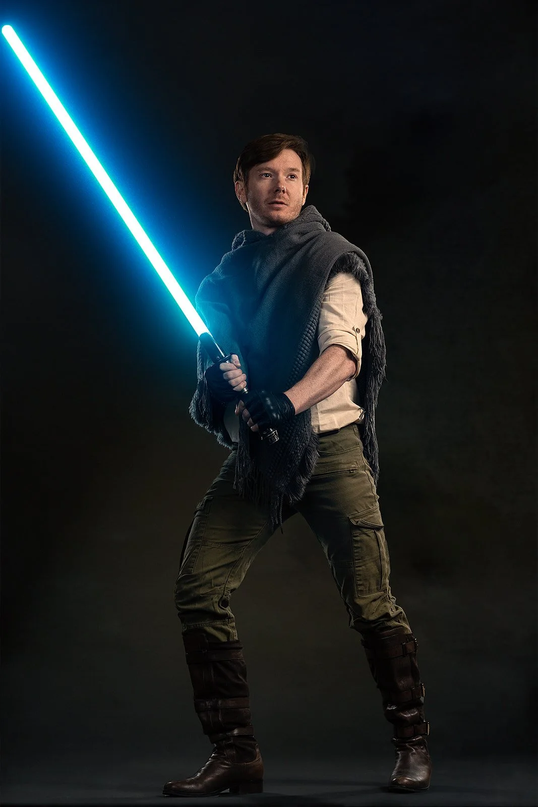 The image is a studio cosplay full body portrait of a white man dressed as Cal Kestis, the protagonist from the Star Wars: Fallen Order game. He embodies the character's essence as he confidently wields a lightsaber, ready for action. His gaze is dir