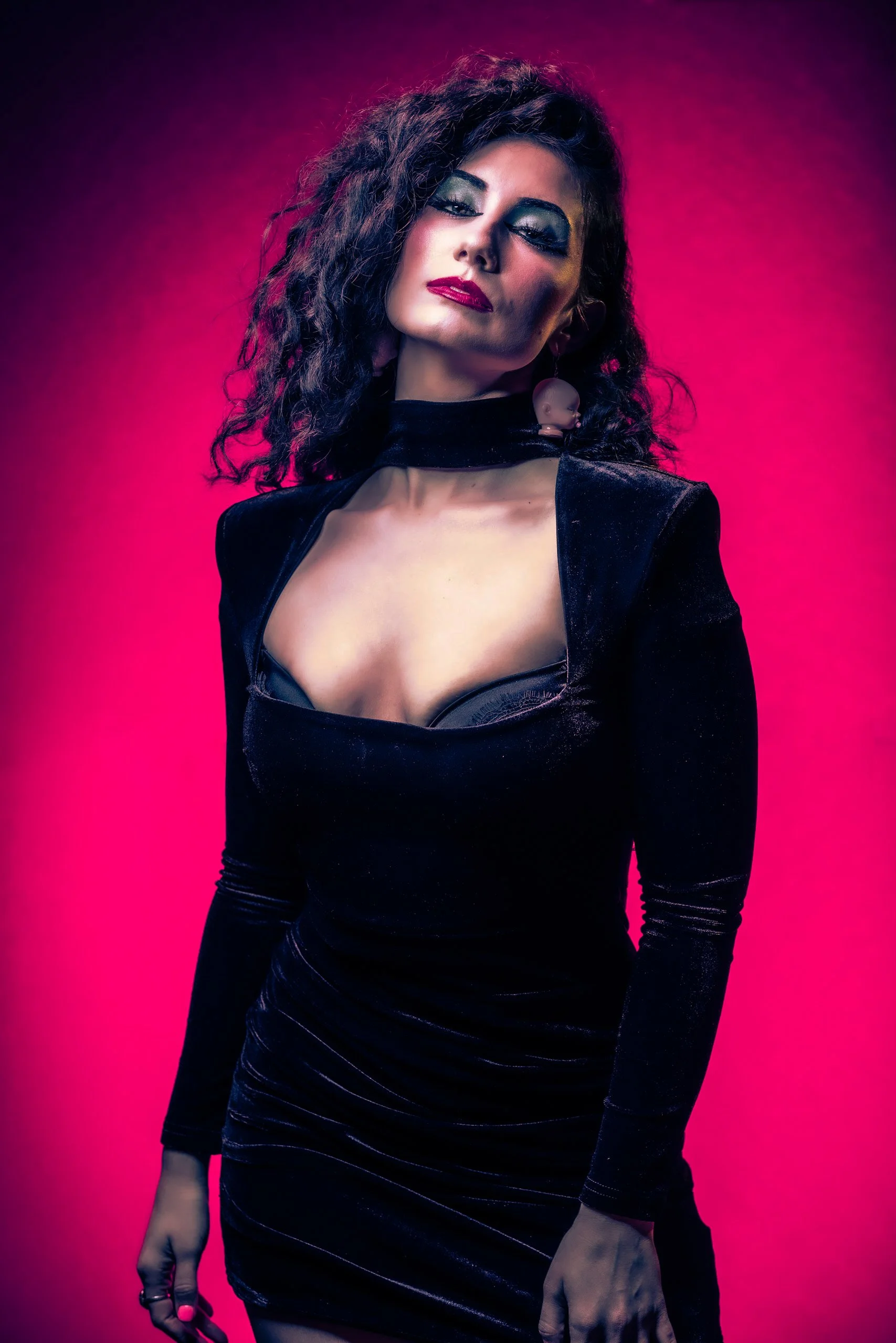 A studio portrait of a white professional model dressed as a 1980's goth. The woman is adorned in a black dress, exuding an edgy and stylish vibe. The image features a fashion aesthetic against a captivating pink background.
