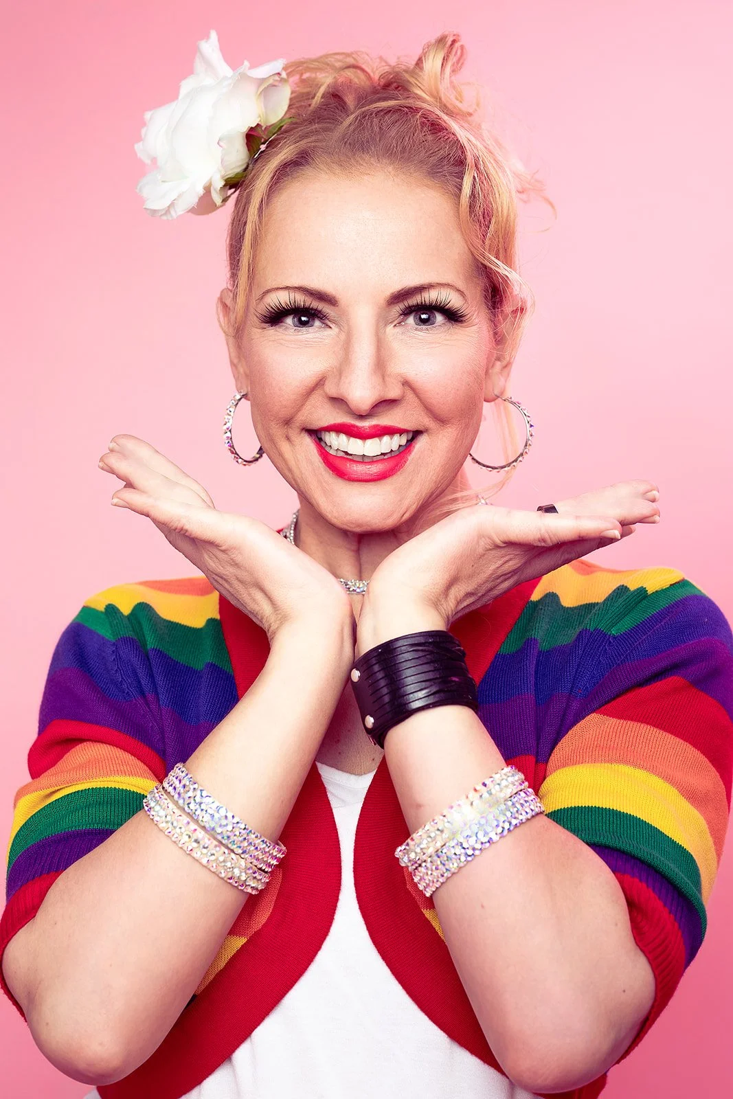 A cheerful woman smiles in a portrait, donned in a vibrant rainbow cardigan. The high-key commercial lighting accentuates her joy, set against a Barbie pink background.