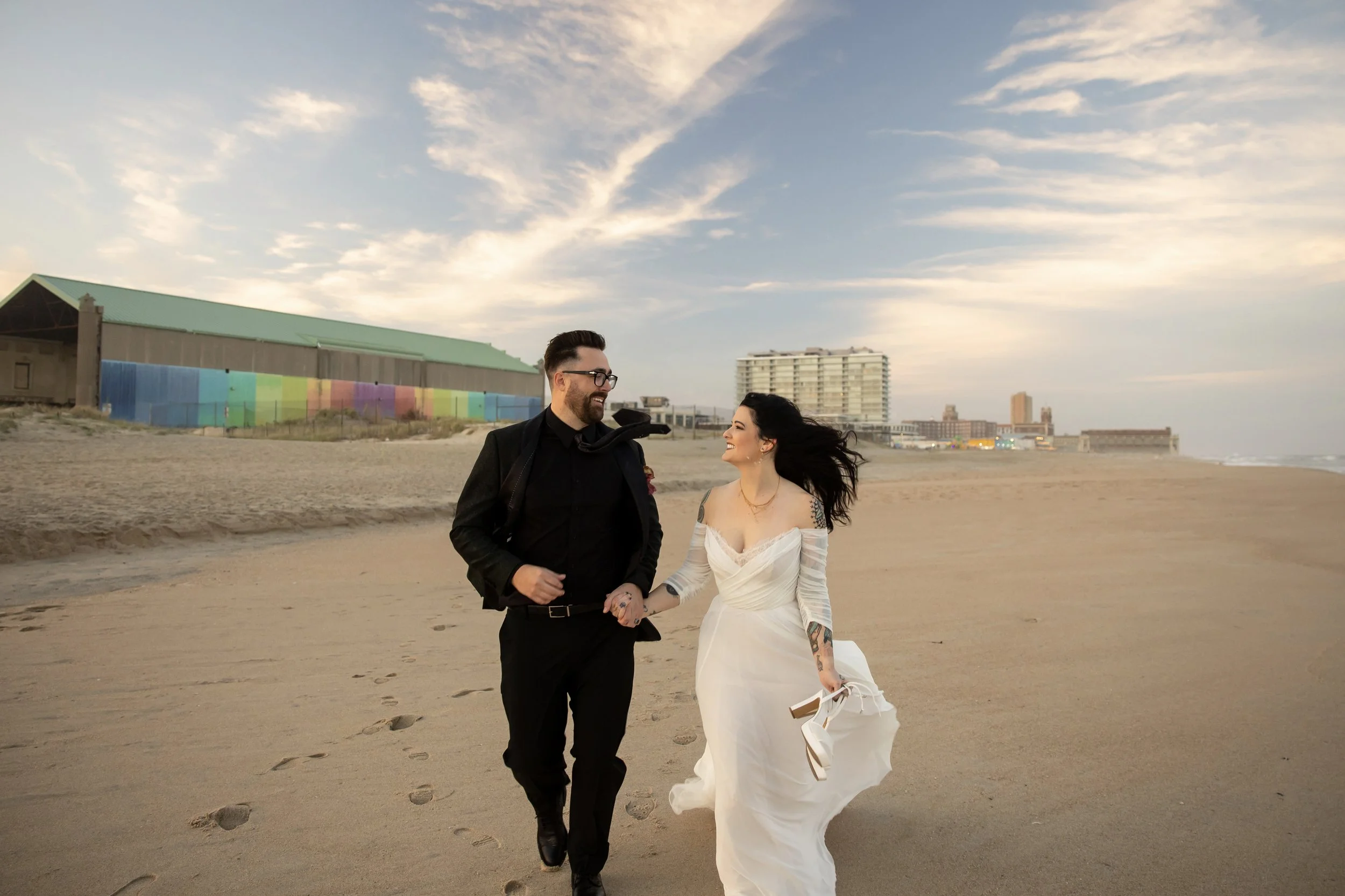 A couple walking on the beach, holding hands and smiling at each other, with a cityscape and colorful building in the background during sunset.