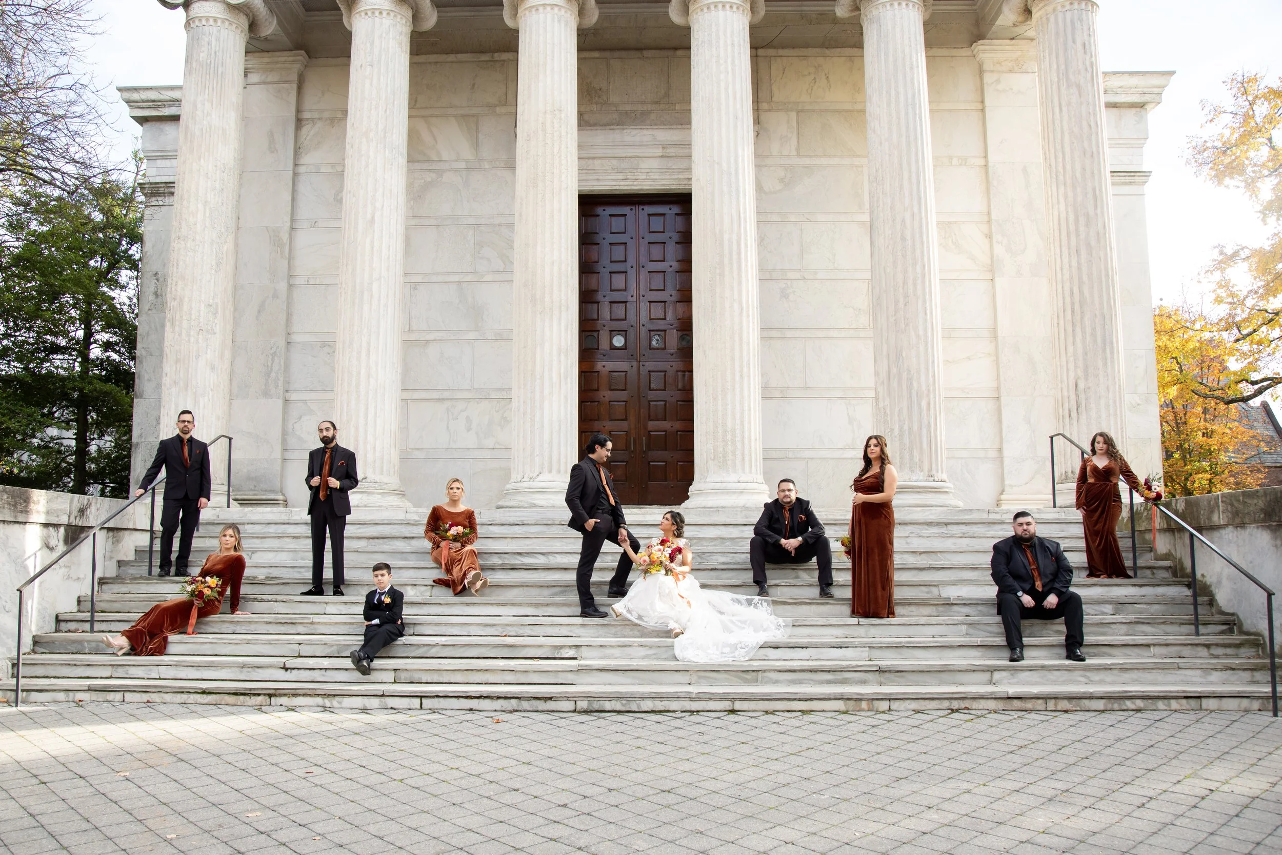 A group of people in formal attire posing on the steps of a large, neoclassical-style building with tall columns and a wooden door, surrounded by trees with autumn foliage.