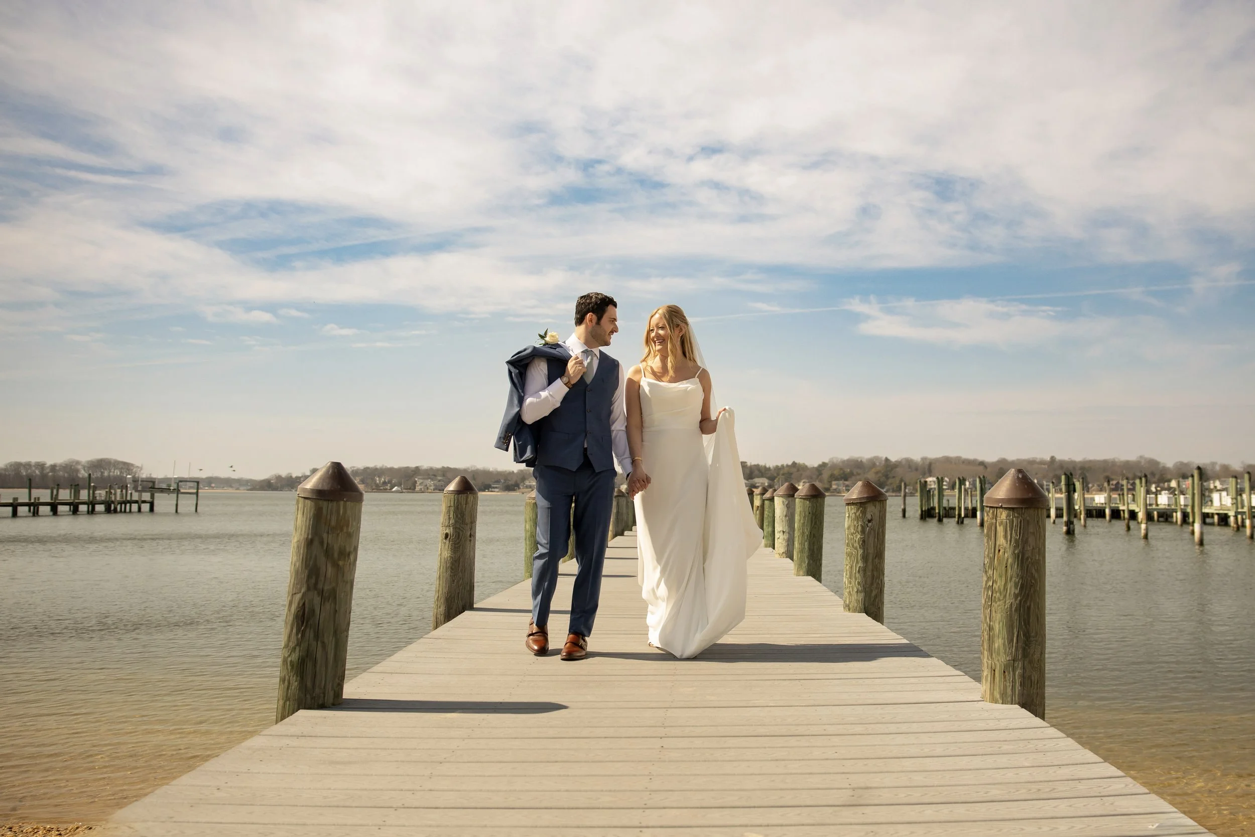 A happy couple walking on a wooden pier by the water, with a partly cloudy sky above. The man is dressed in a suit and is carrying a jacket over his shoulder, while the woman is wearing a white dress. They are holding hands and smiling at each other.
