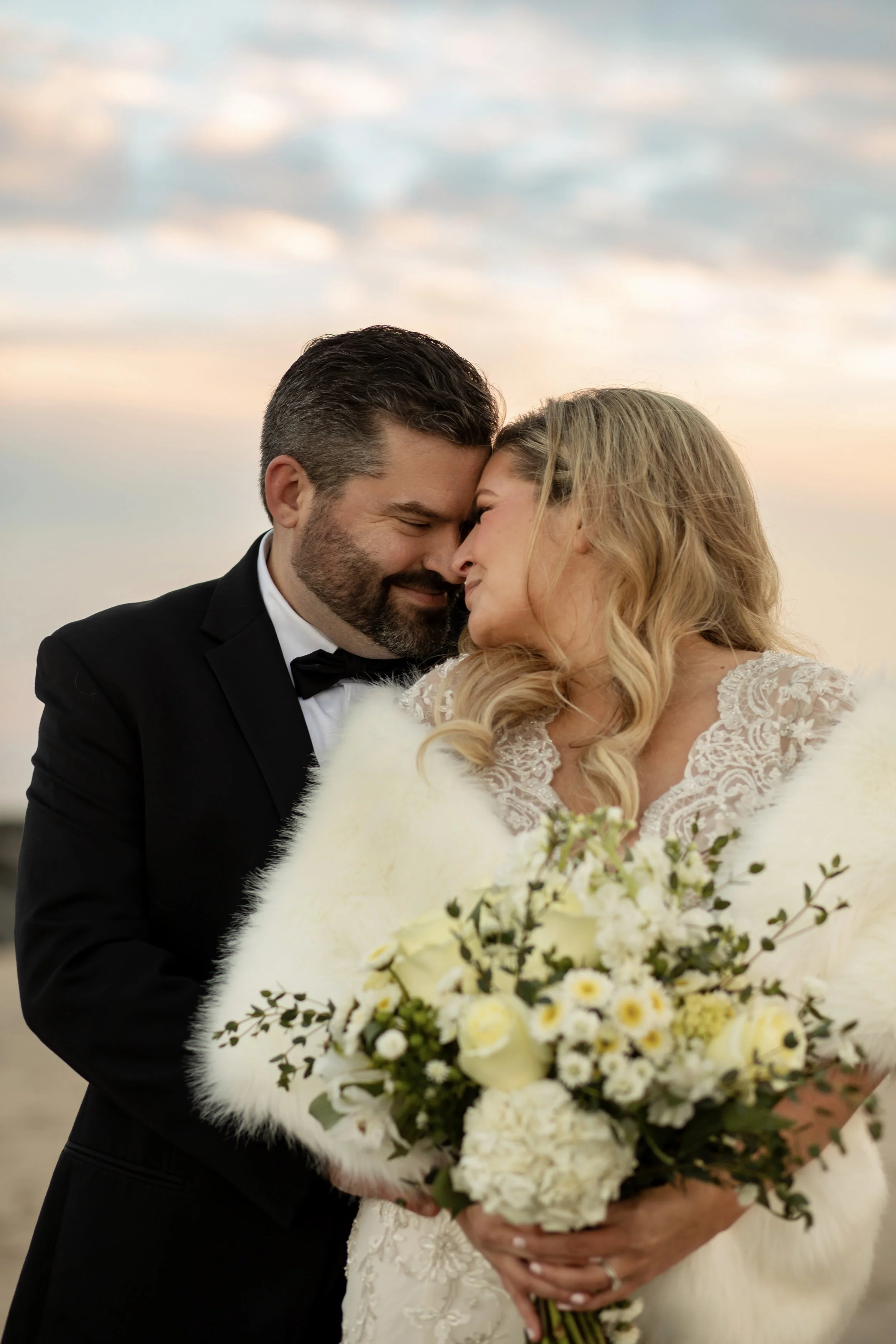 A bride and groom share a close moment during their wedding, with the bride holding a large bouquet of white and yellow flowers and they are touching foreheads, set against a soft sky background.