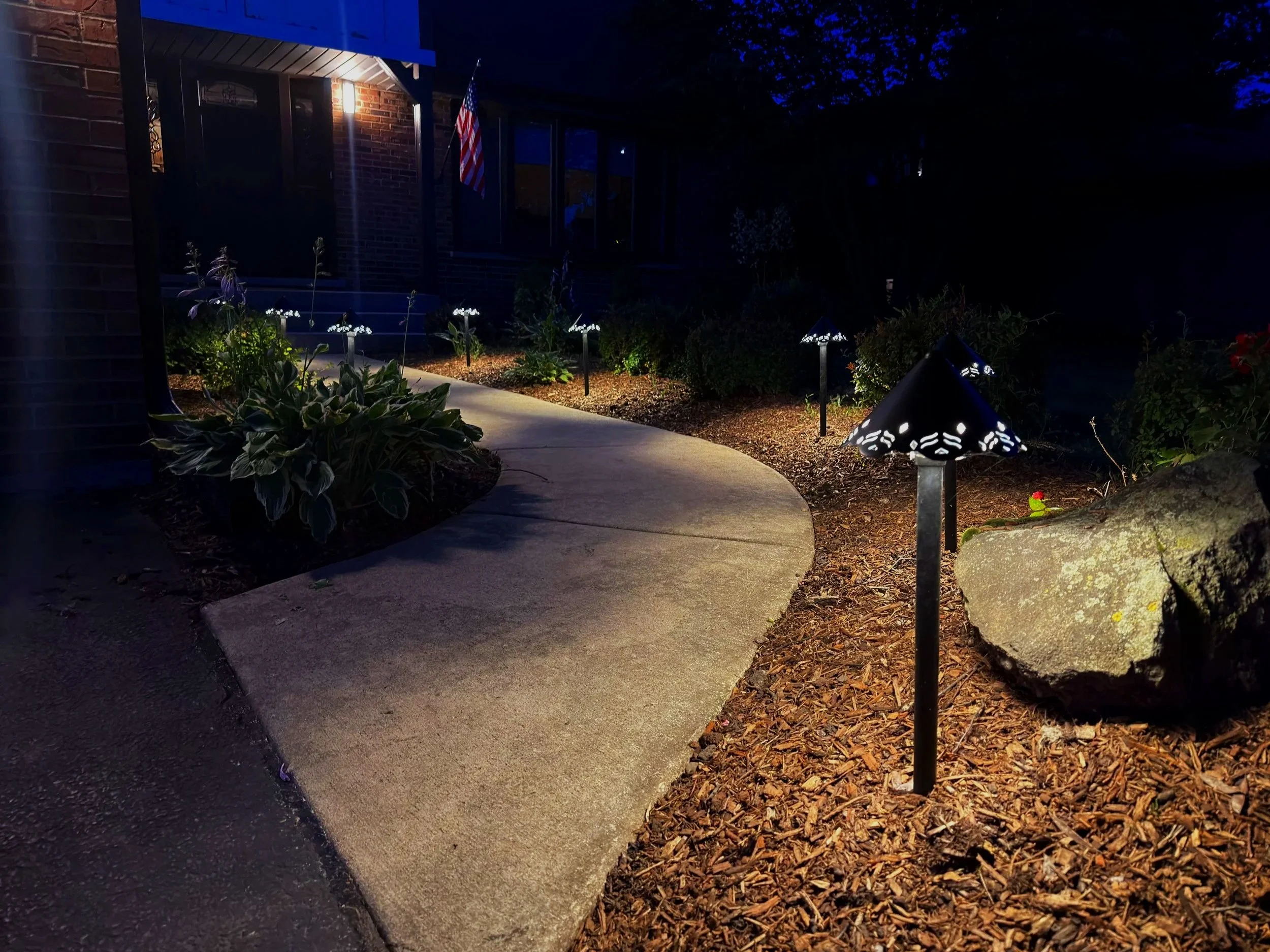Wide night view of a curved concrete walkway, path lights, and mulched planting beds beside a brick residence.