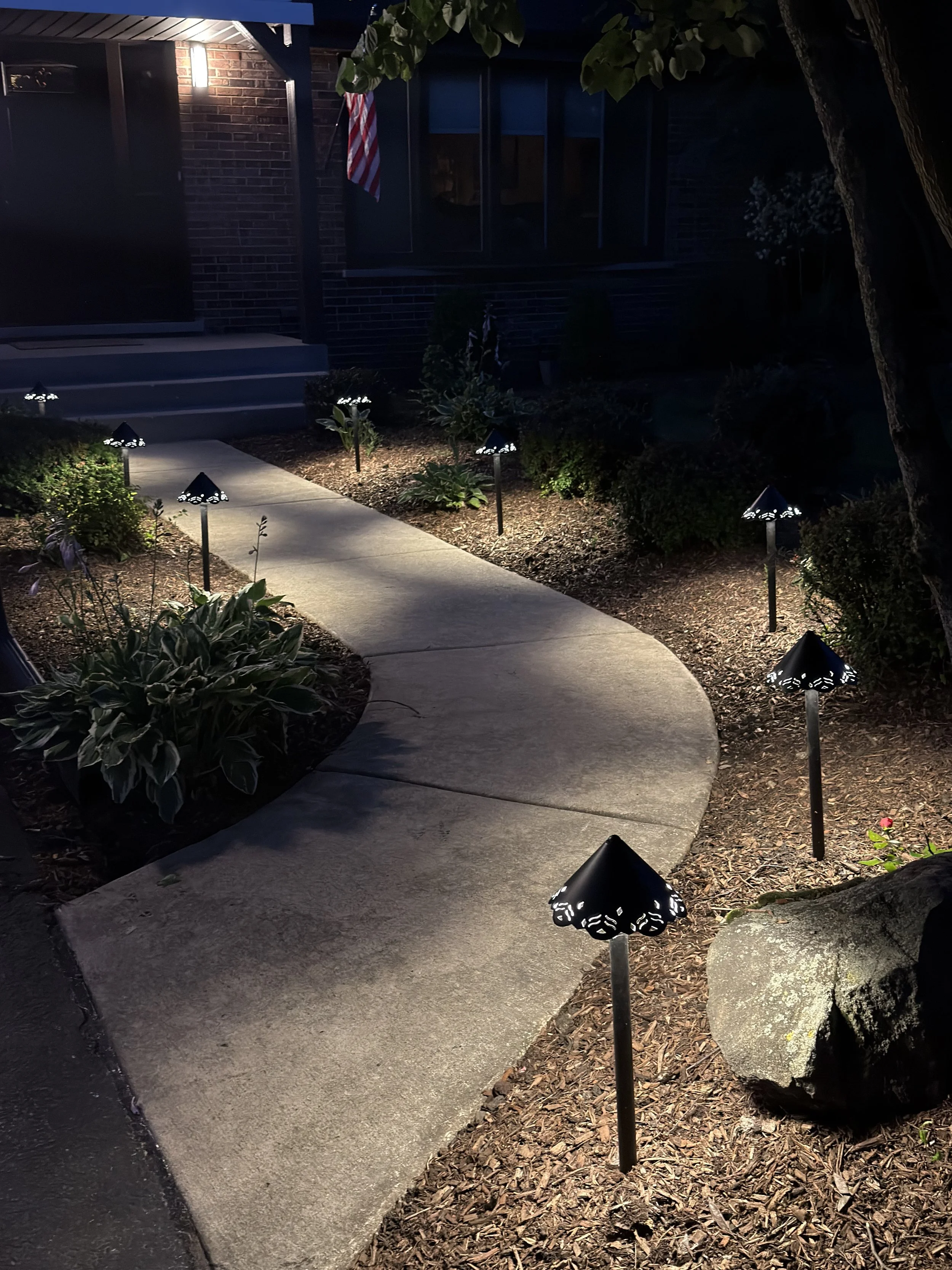 Night view of a curved concrete walkway with black path lights, decorative stone, and planting beds beside a brick home.