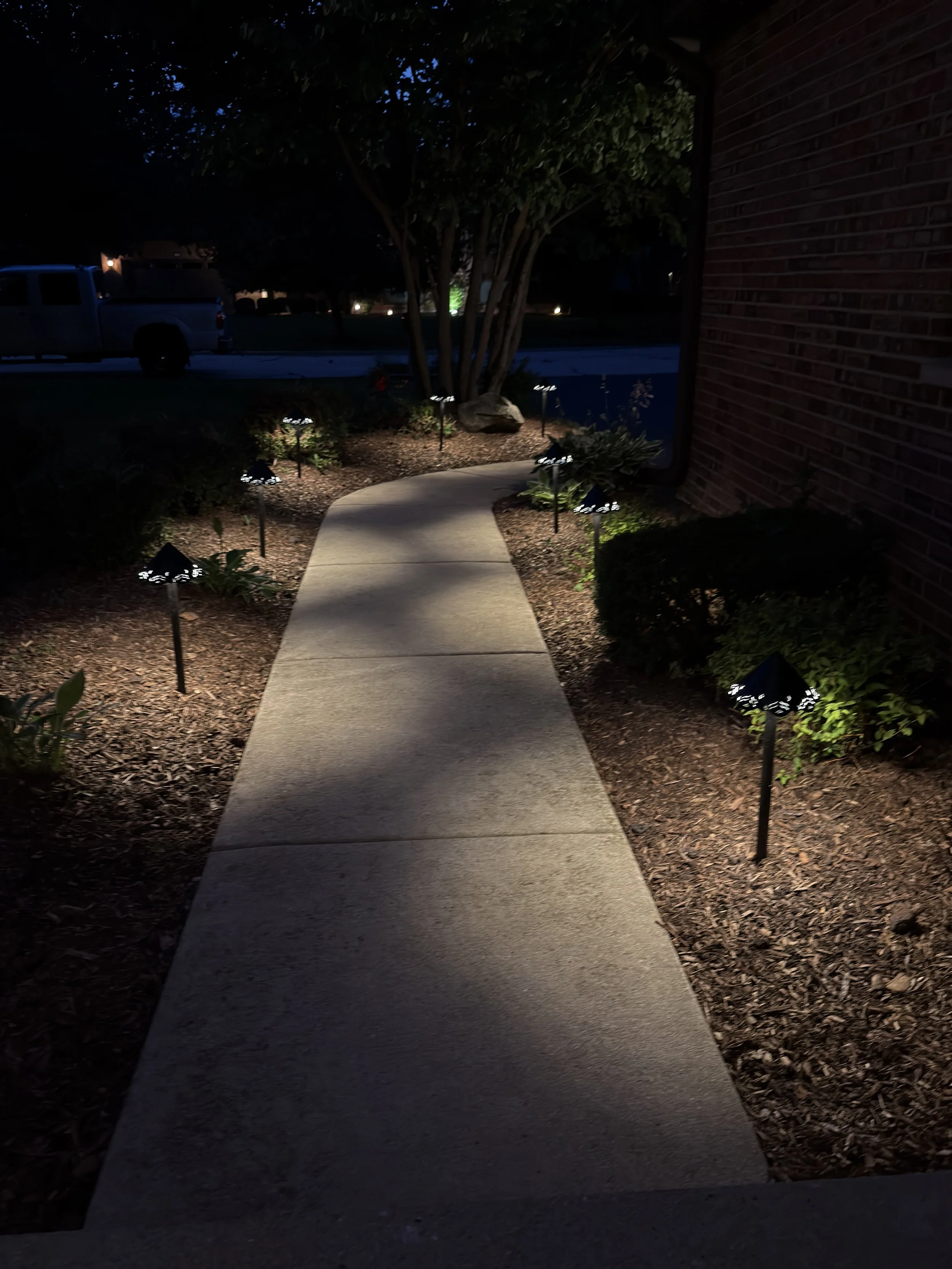 Straight-on night view of an illuminated concrete walkway bordered by path lights, shrubs, and a brick wall.