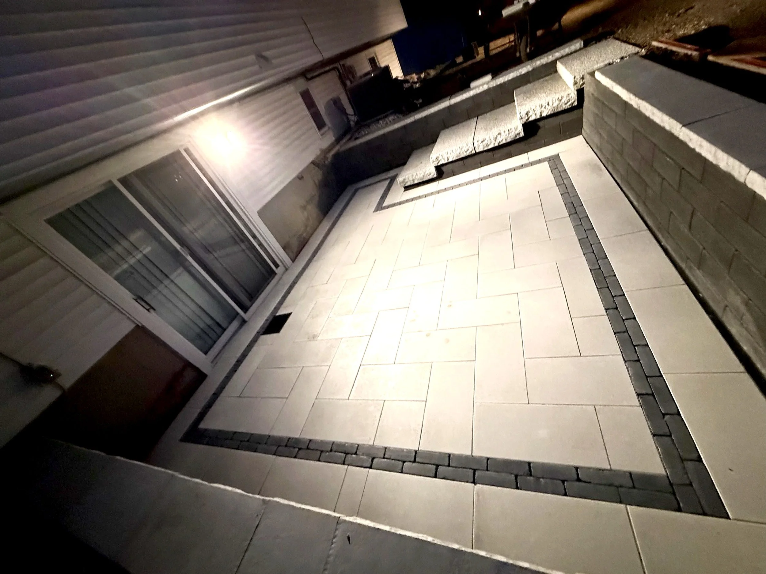 An outdoor space at night with a tiled patio, a brick half wall, a window with a white frame, and a house with horizontal siding illuminated by a wall-mounted light fixture.