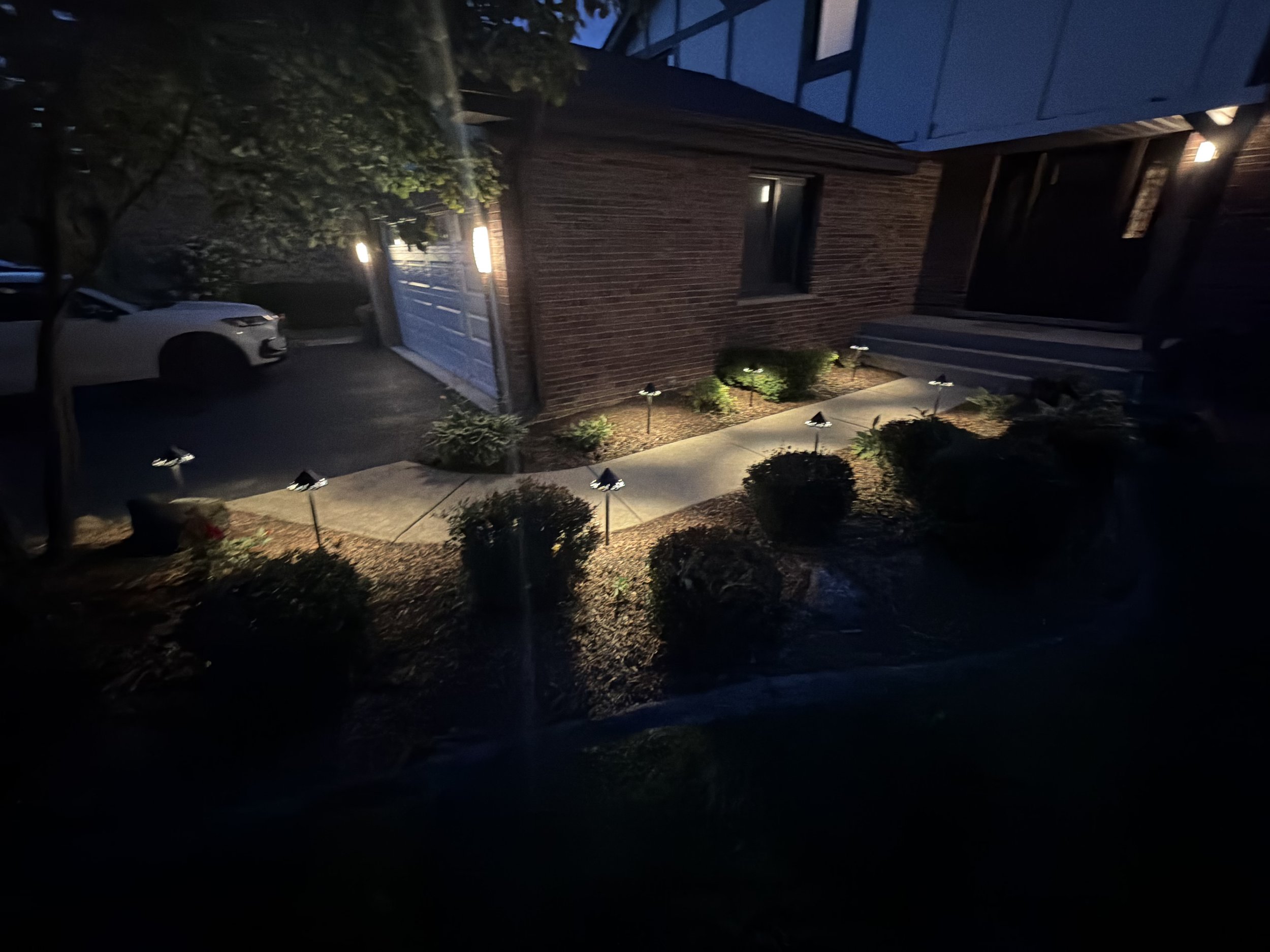 Night view of a brick home, driveway edge, shrubs, and landscape lights near the garage.
