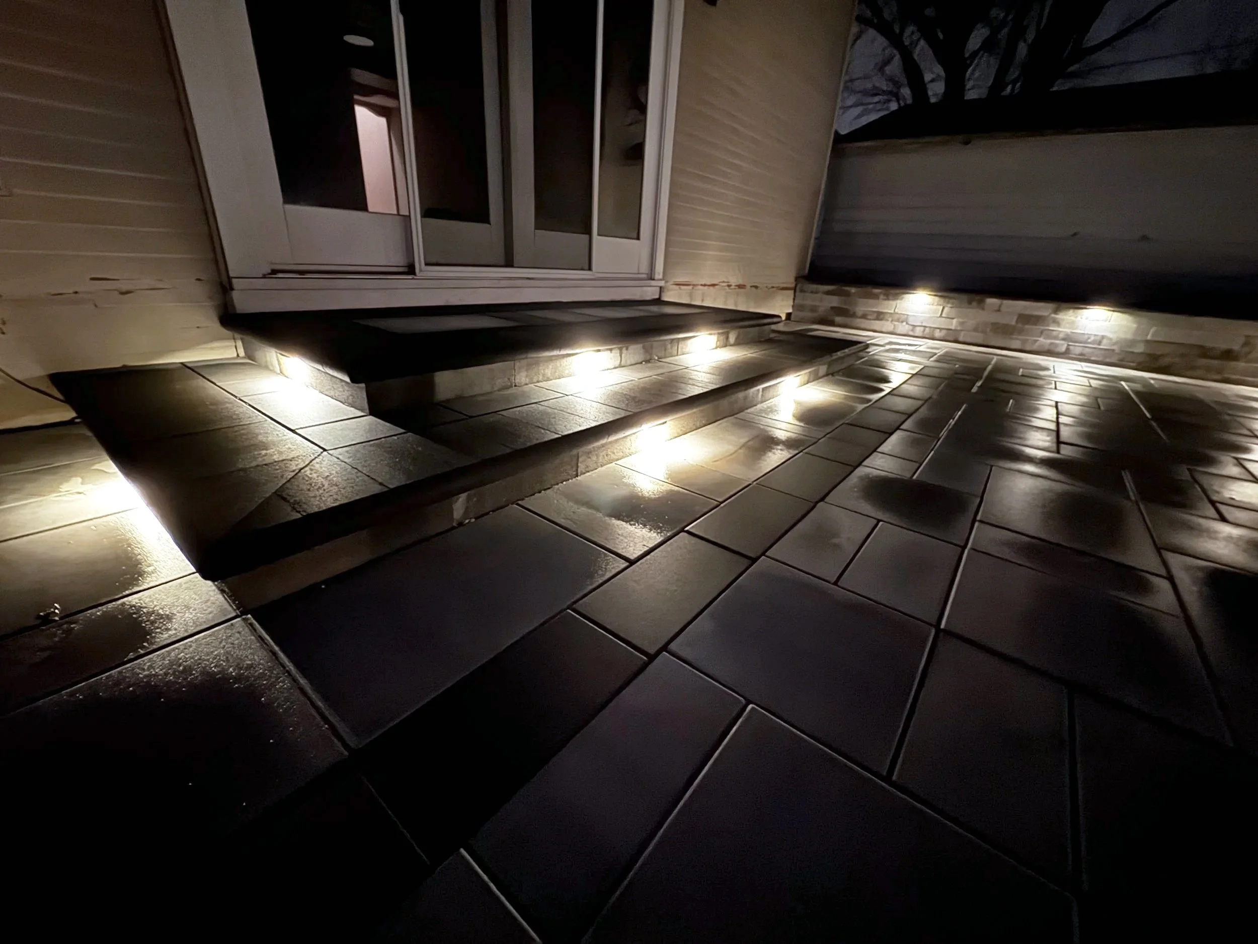Nighttime porch scene with illuminated steps leading to a sliding glass door on a house with wood siding, a tree visible in the background.
