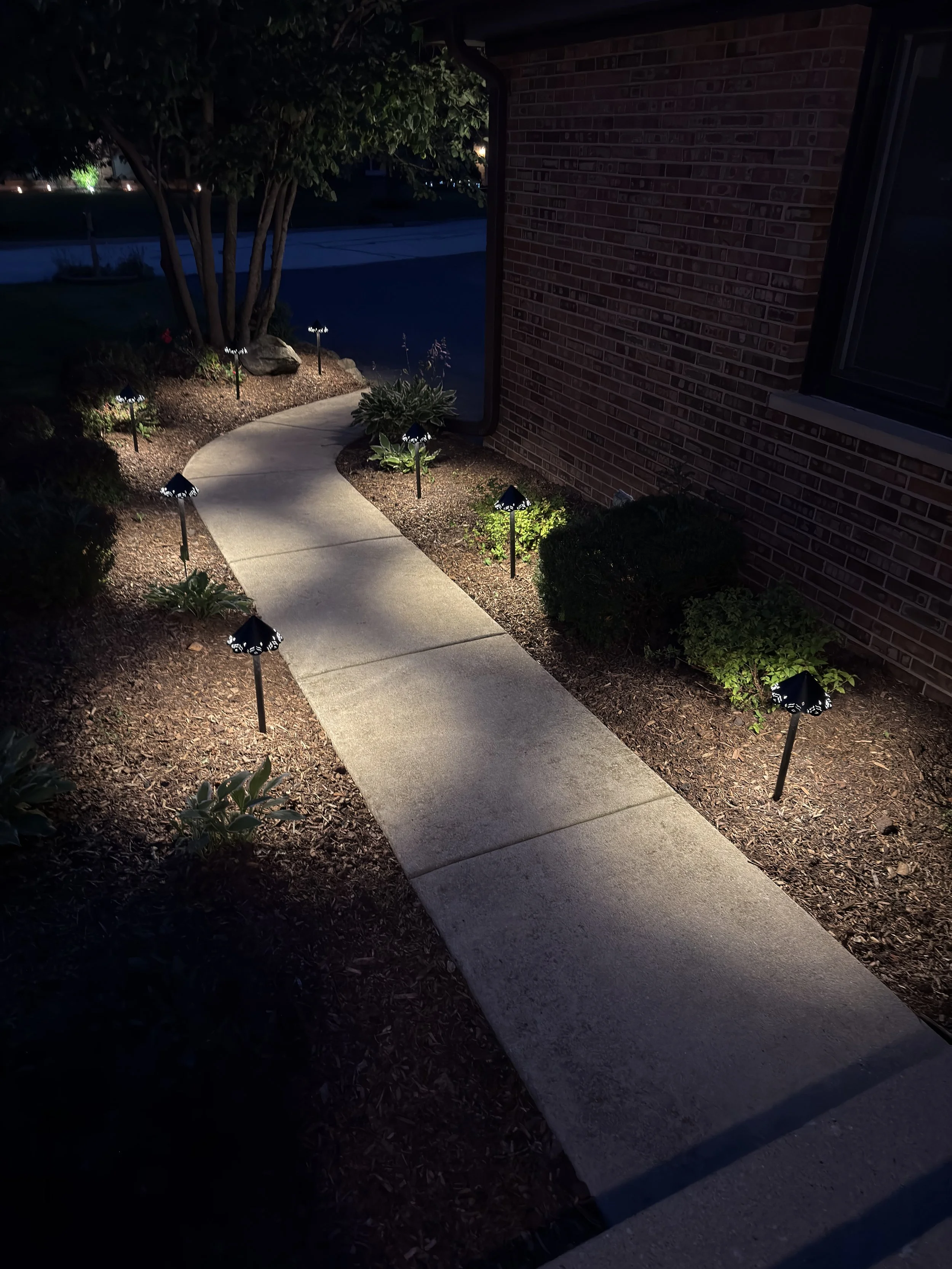 Vertical night view of a straight concrete walkway illuminated beside a brick home and mature tree.