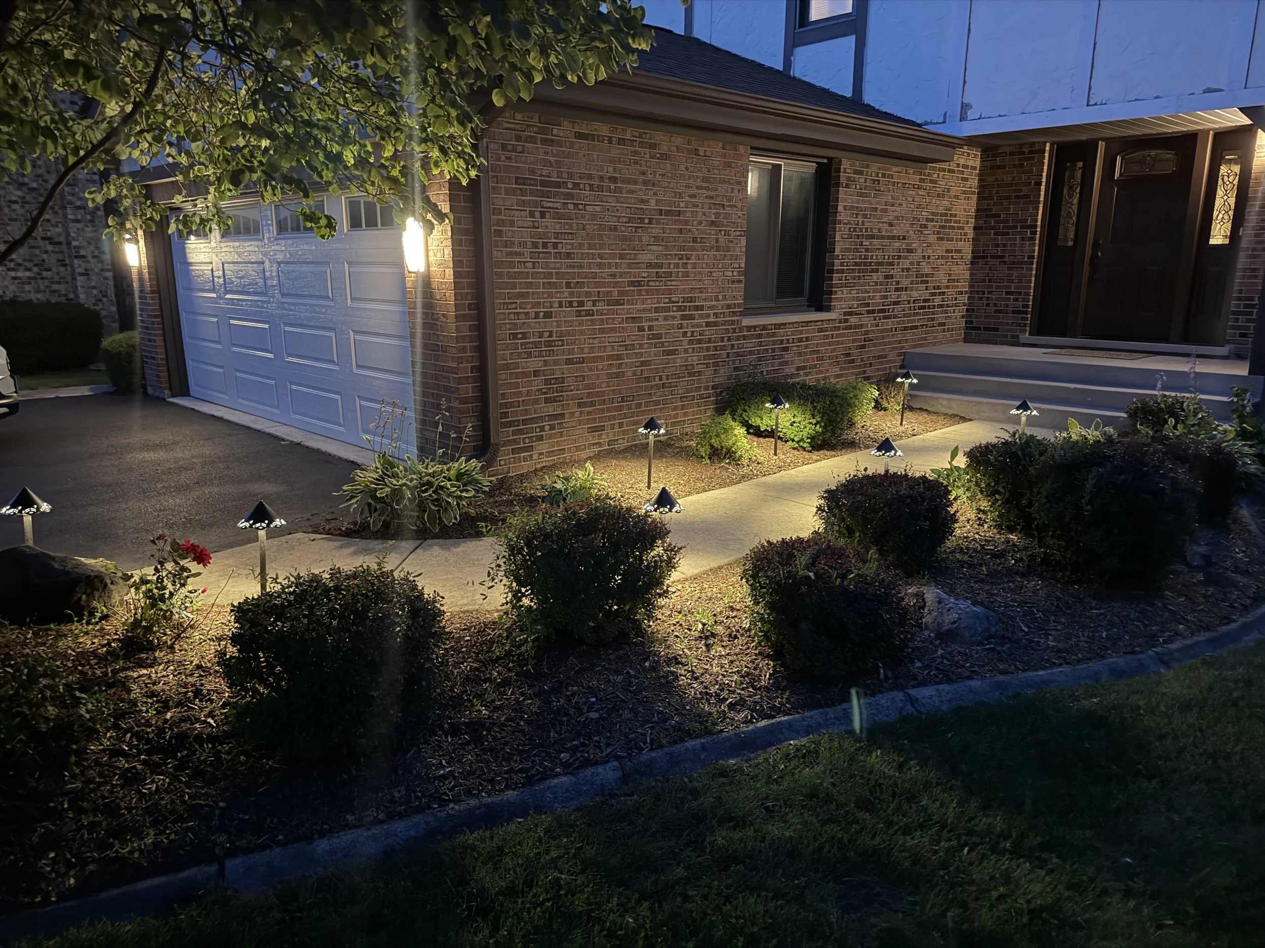 Wide dusk view of a brick home and garage with accent lighting across the front planting beds.