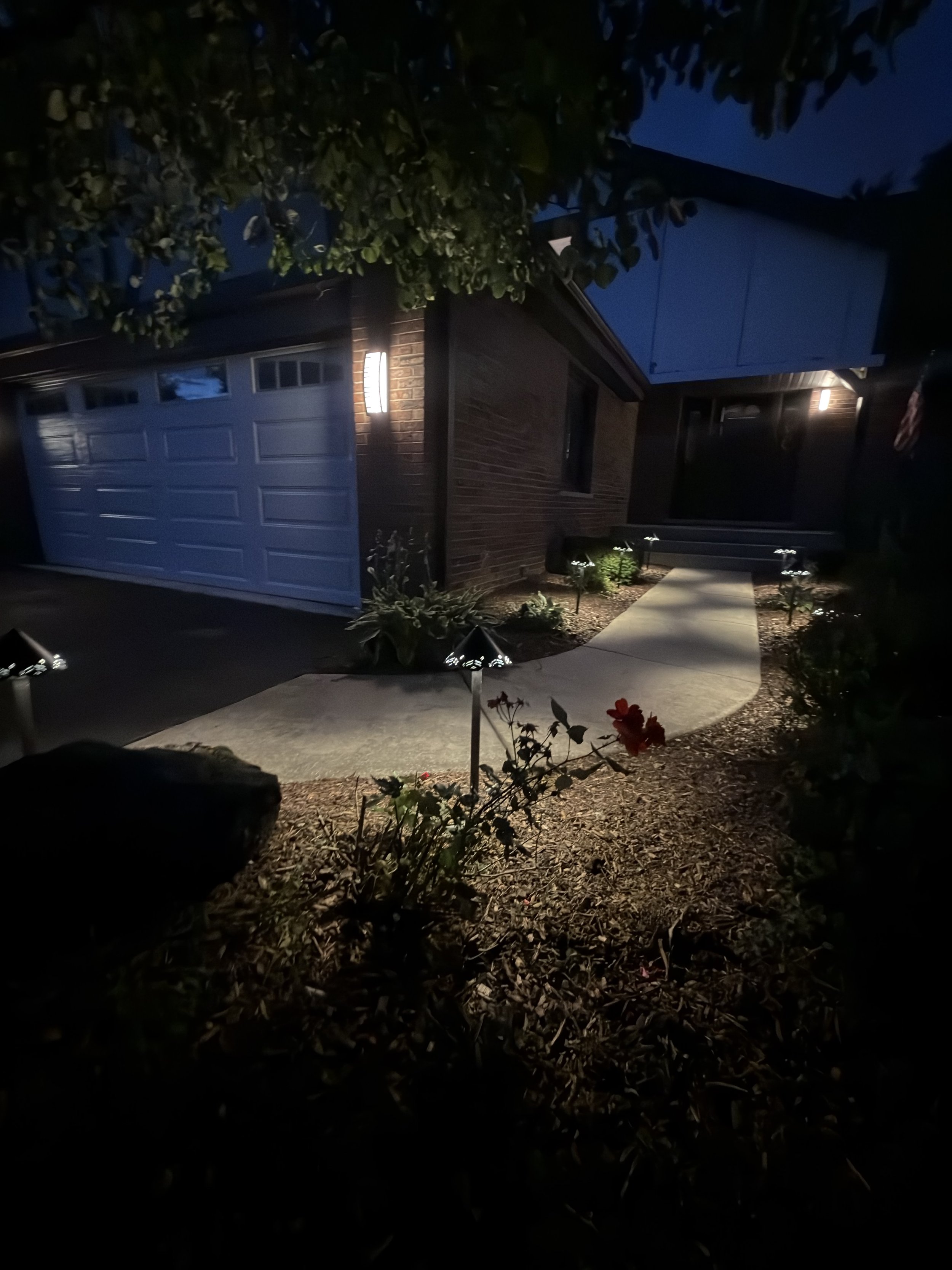 Night view across a landscaped front yard toward a lit walkway and garage beside a brick home.