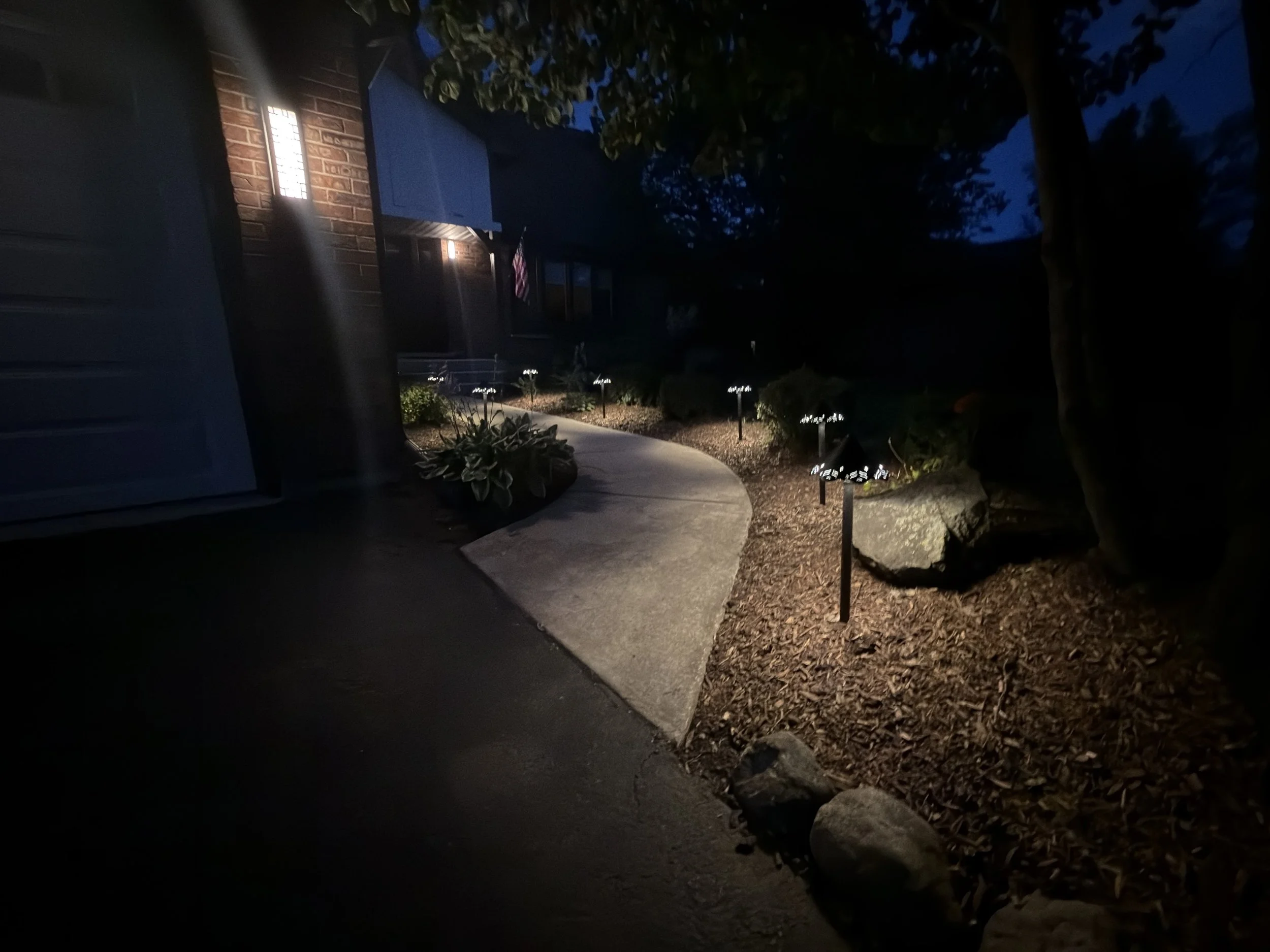 Nighttime view of a curved concrete walkway illuminated by black path lights beside a brick home and mulched garden beds.