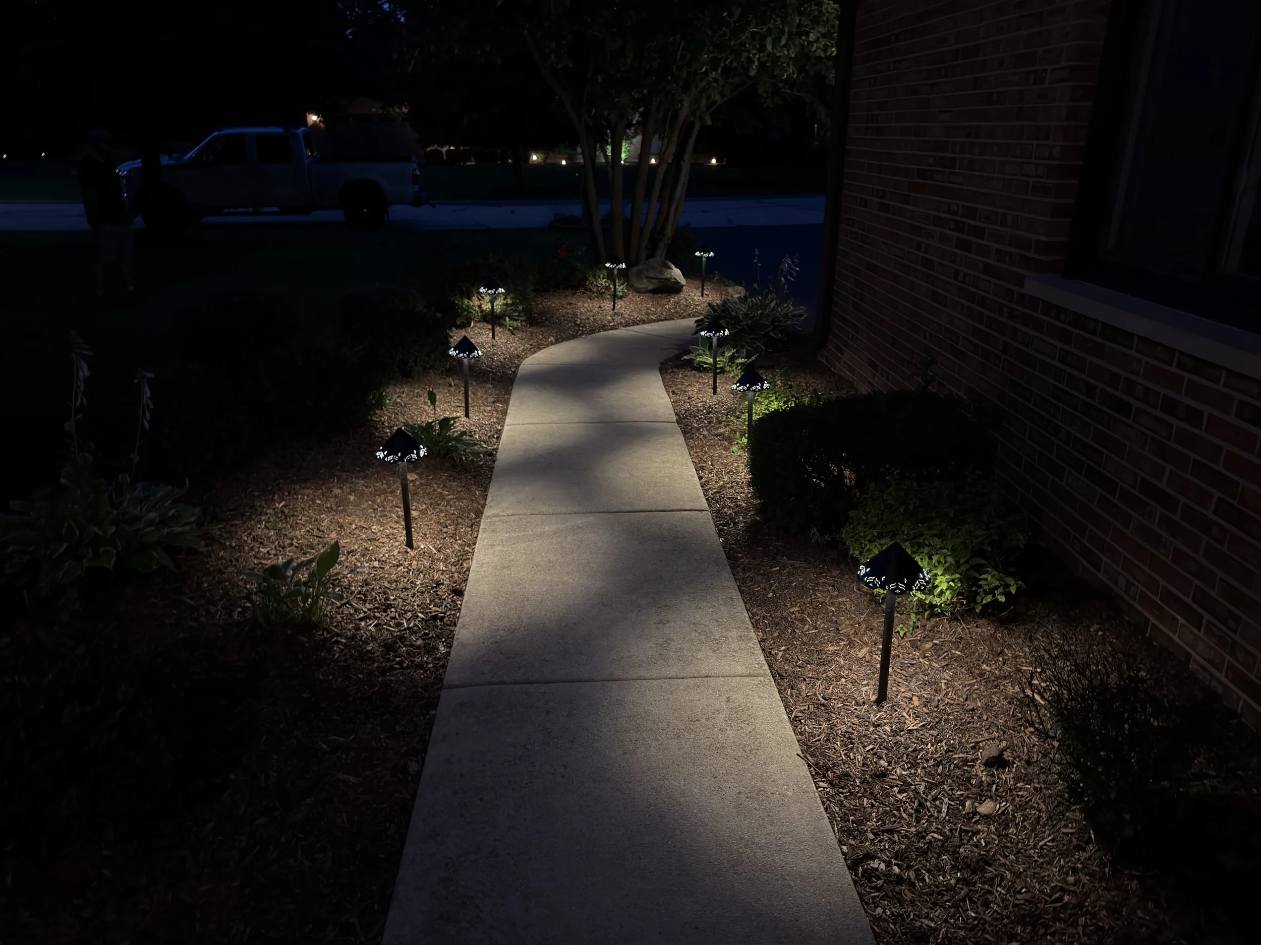 Wide night view of a lit walkway and landscaped bed leading toward the front of a brick home.