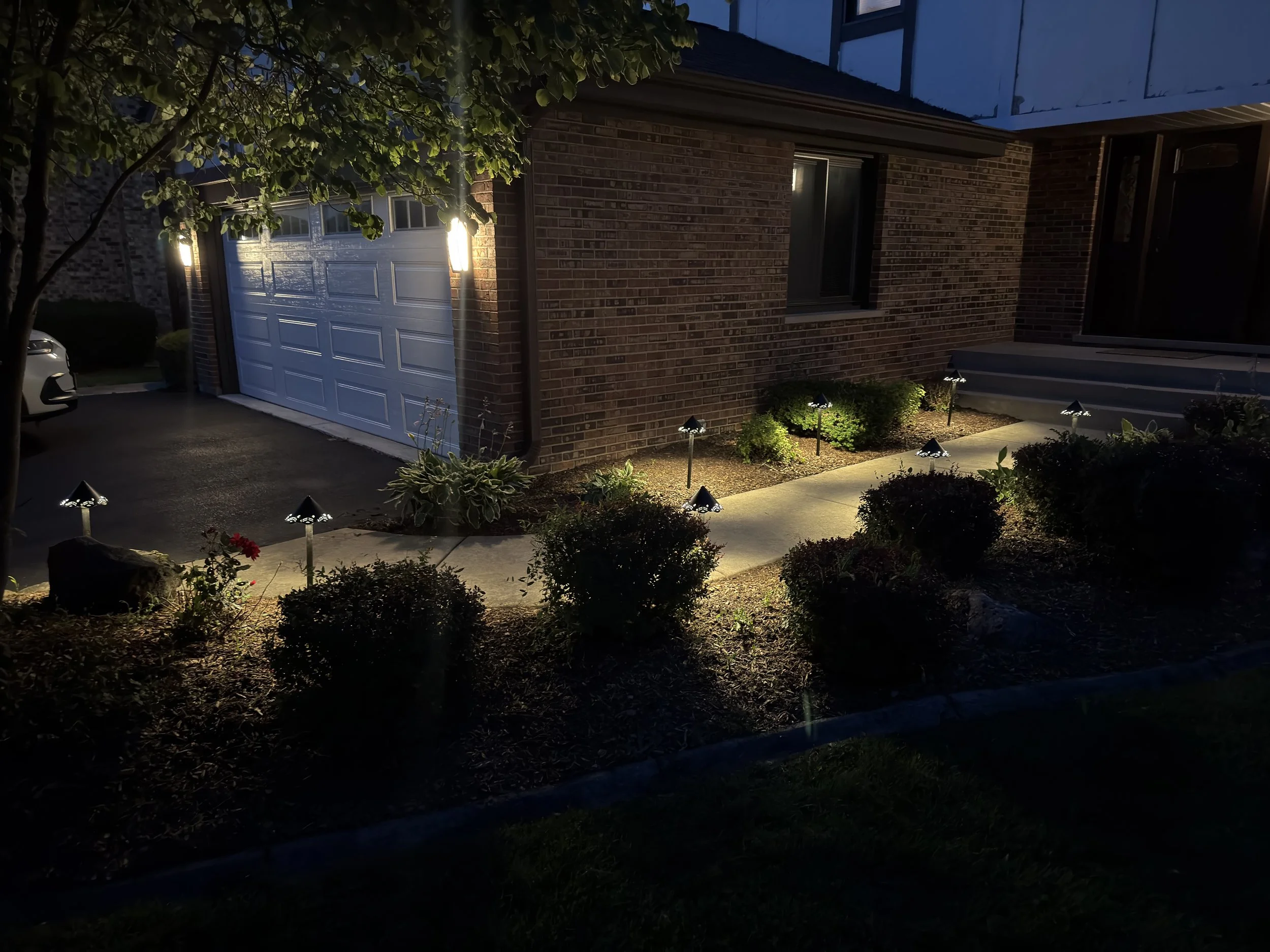 Dusk view of a brick residence with path lights illuminating shrubs, lawn edges, and the walkway.