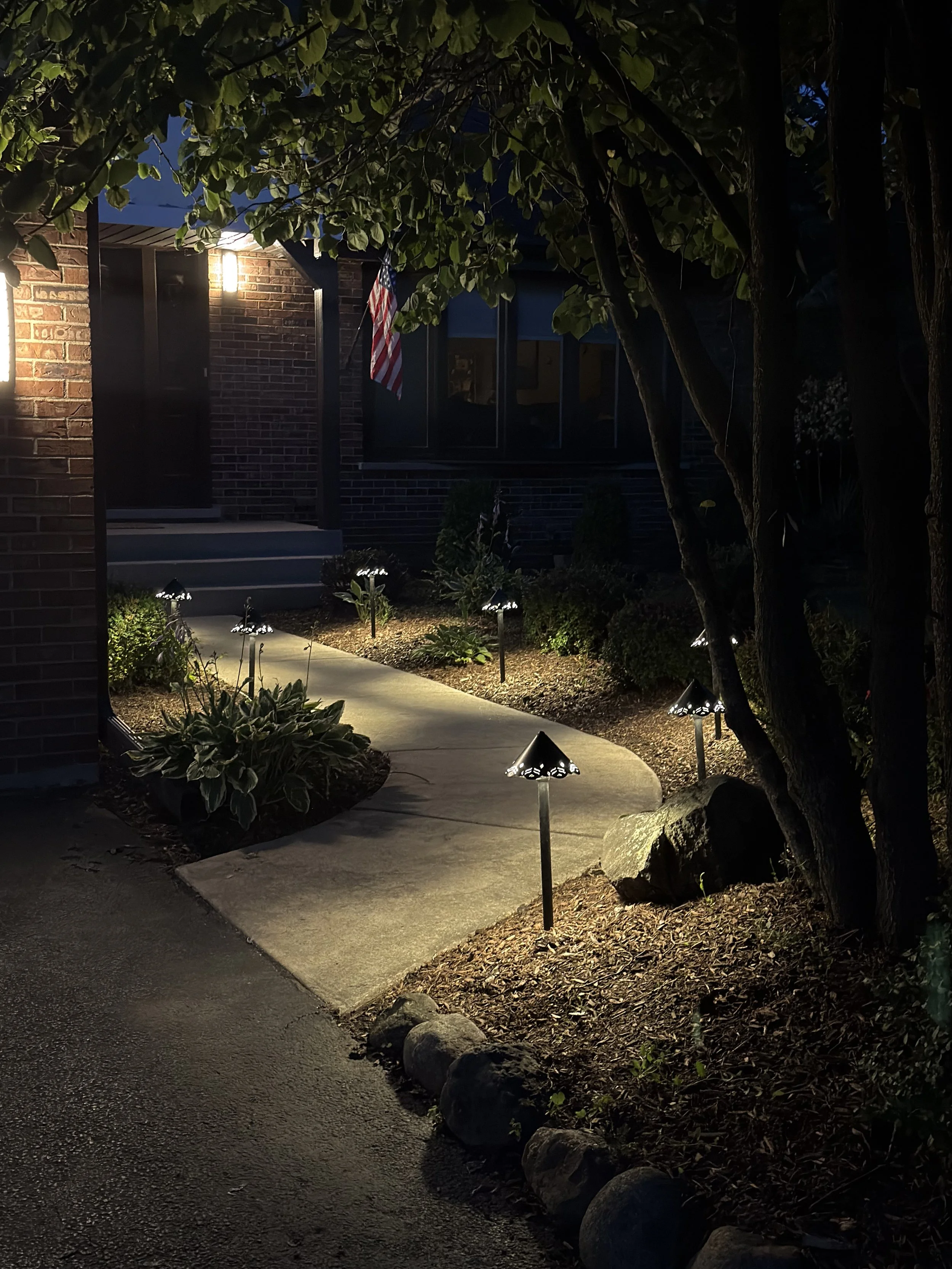 Vertical night view of a curved concrete walkway with a bright path light, boulders, and shaded trees.
