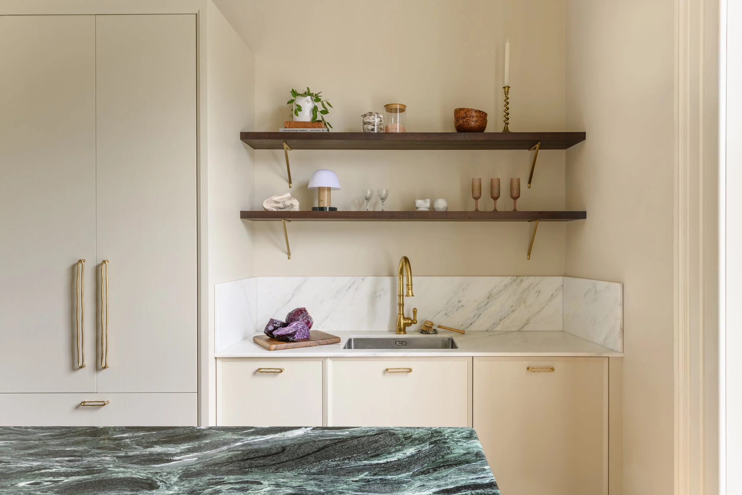 Kitchen with cream cabinets, marble backsplash, golden faucet, open wooden shelves with decorative items, purple stones on a wooden cutting board, and marble countertop in the foreground.