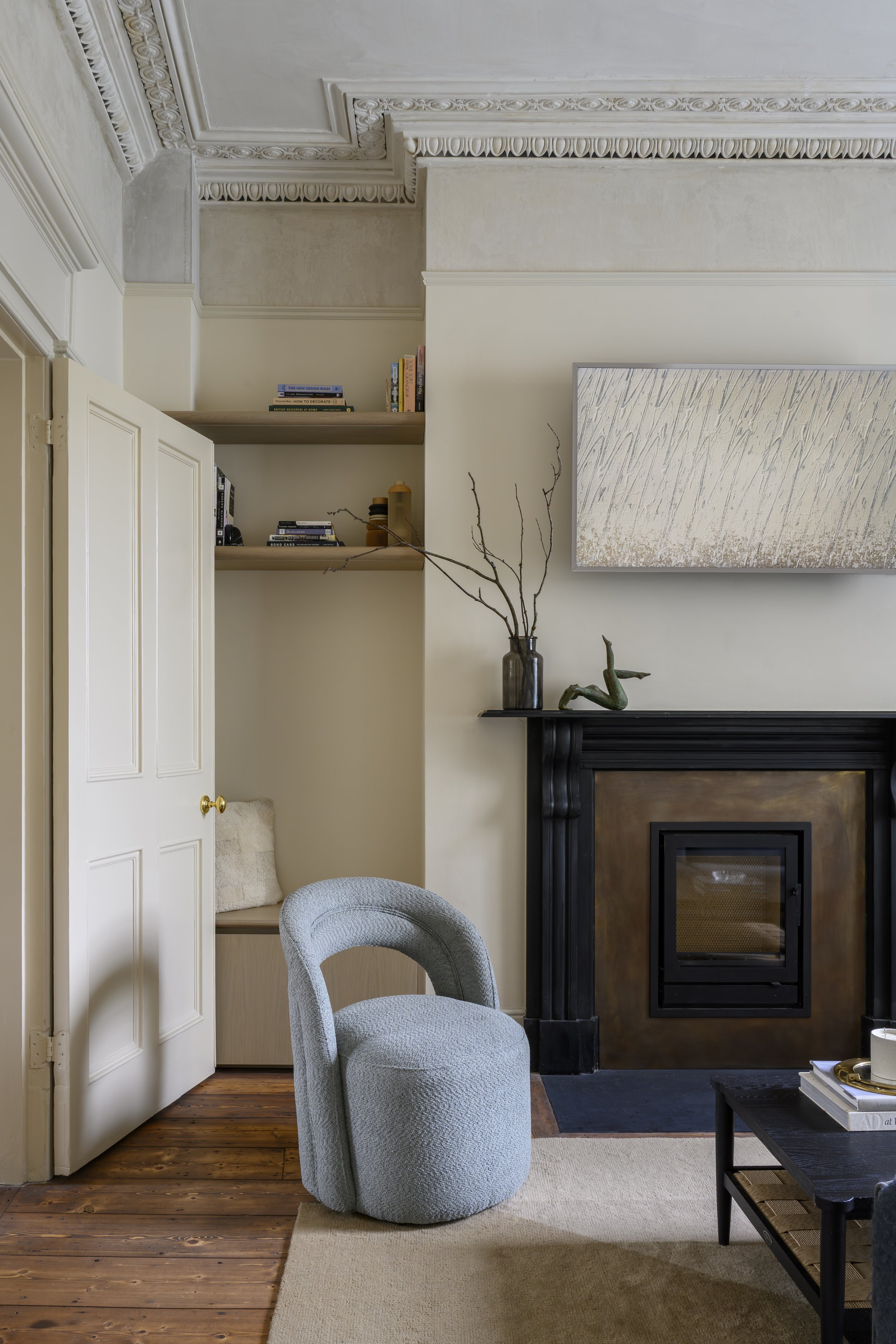 A living room with a gray upholstered chair in front of a fireplace, a beige rug, a black side table with books and a white mug, a cream closet with open shelves containing books and decorations, a branch in a black vase, and modern wall art above the fireplace.