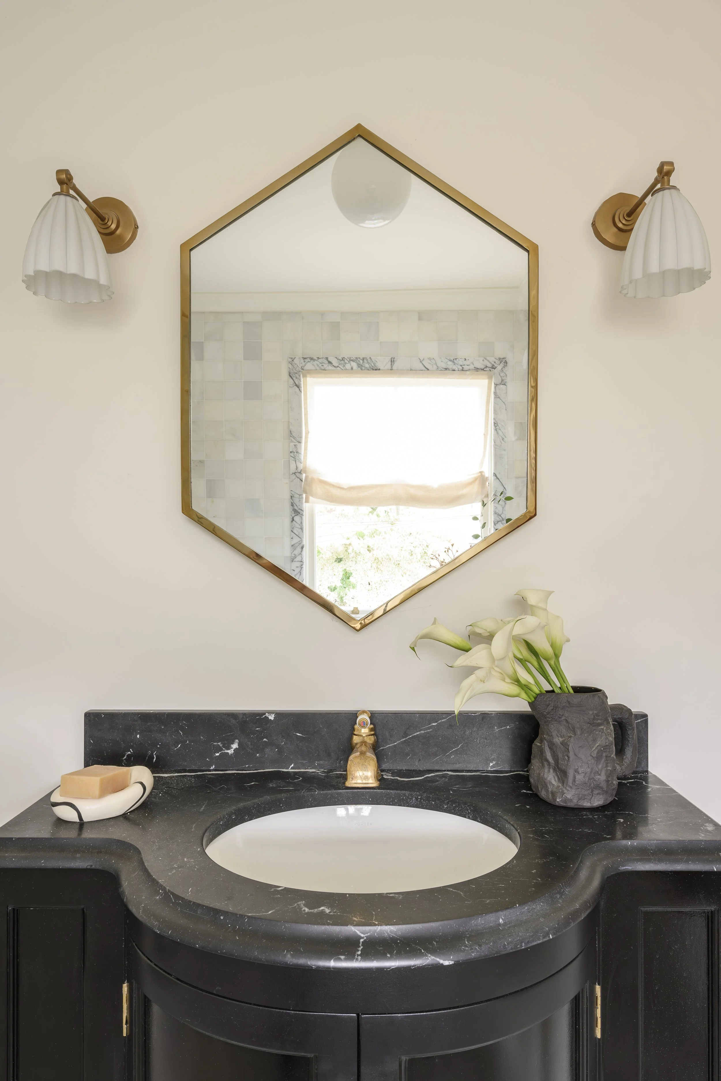 Bathroom vanity with a black marble countertop, white sink, gold faucet, a vase with white calla lilies, a soap dish with soap, a mirror, and two wall-mounted light fixtures.
