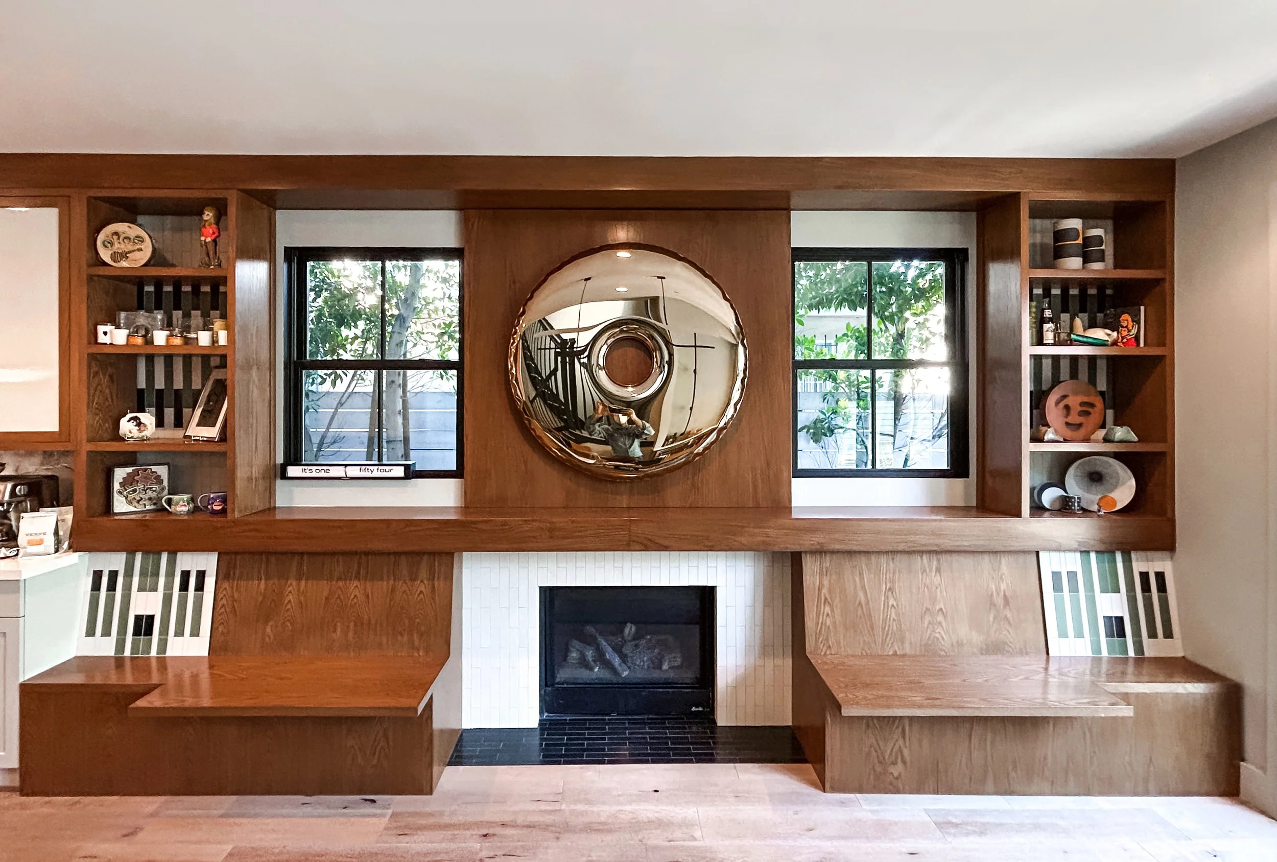 Interior with modern wooden shelving, a large circular mirror, two windows, and a central fireplace. Light walls and wood flooring complete the minimalist design.