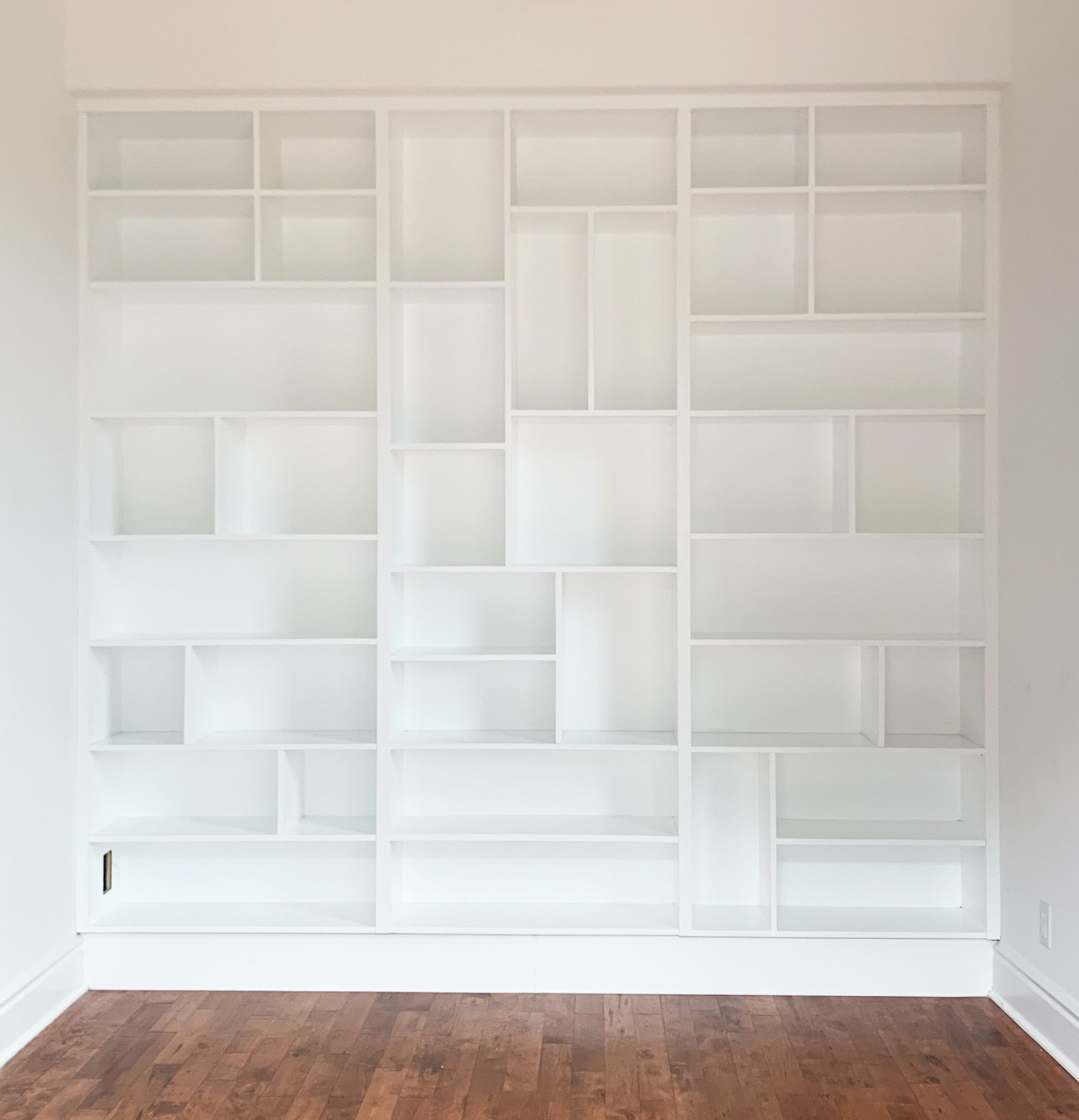 Modern white built-in bookshelf with various-sized compartments against a white wall, above a wooden floor.