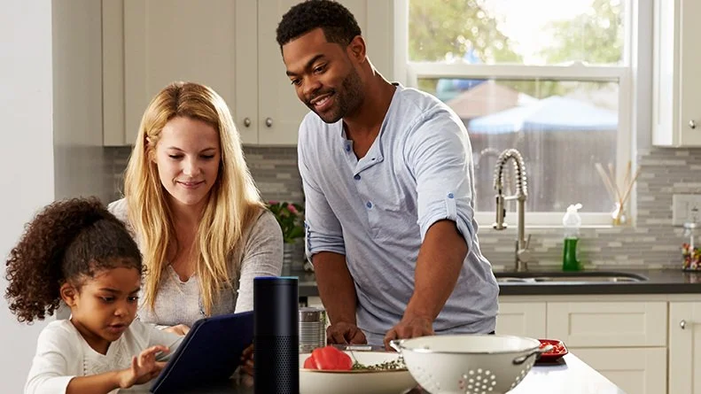 A couple in their kitchen speak to Alexa via the Echo speaker on their counter.