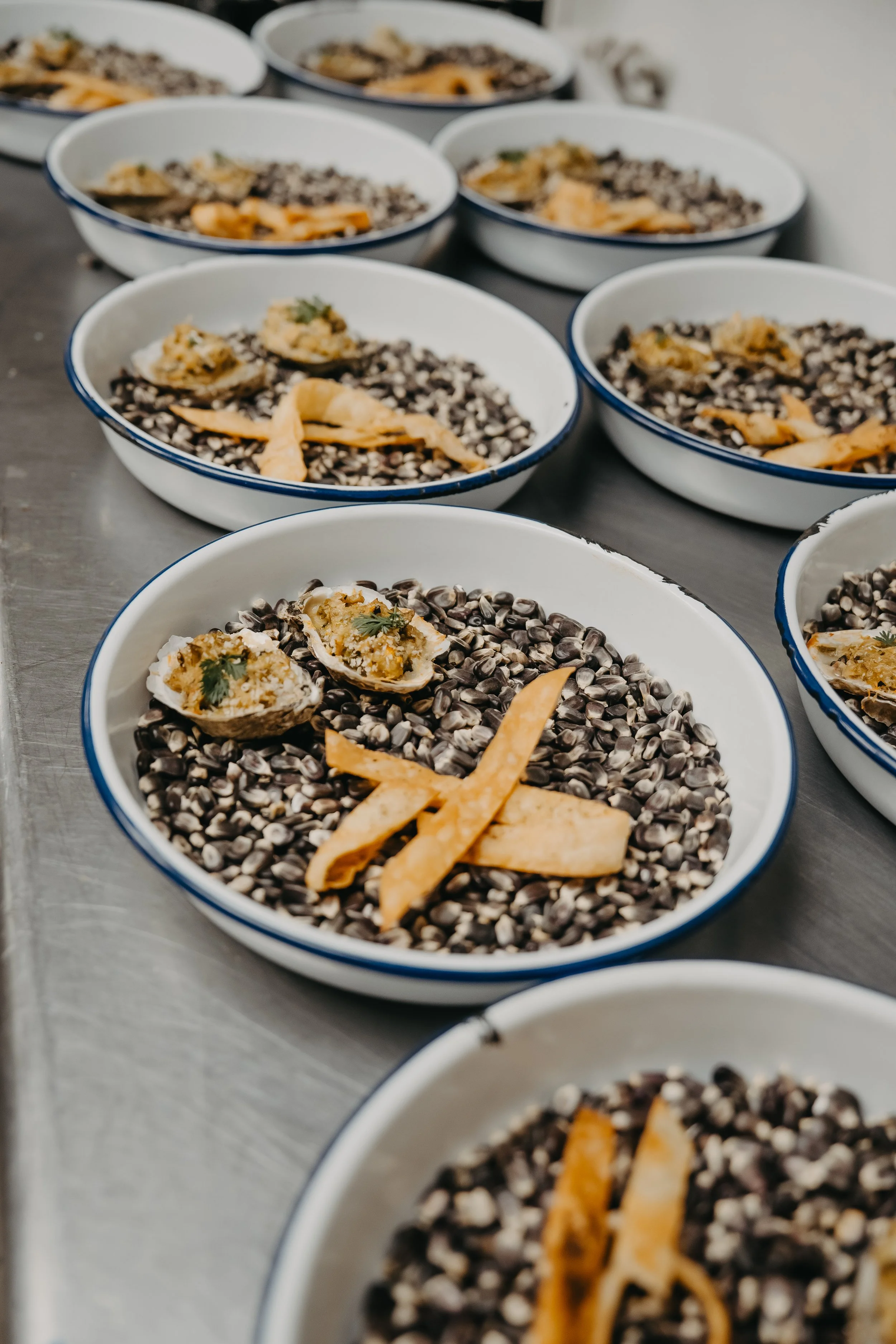Several white bowls filled with black beans, topped with garnishes like sliced tamarind or plantain chips, placed on a metallic surface.