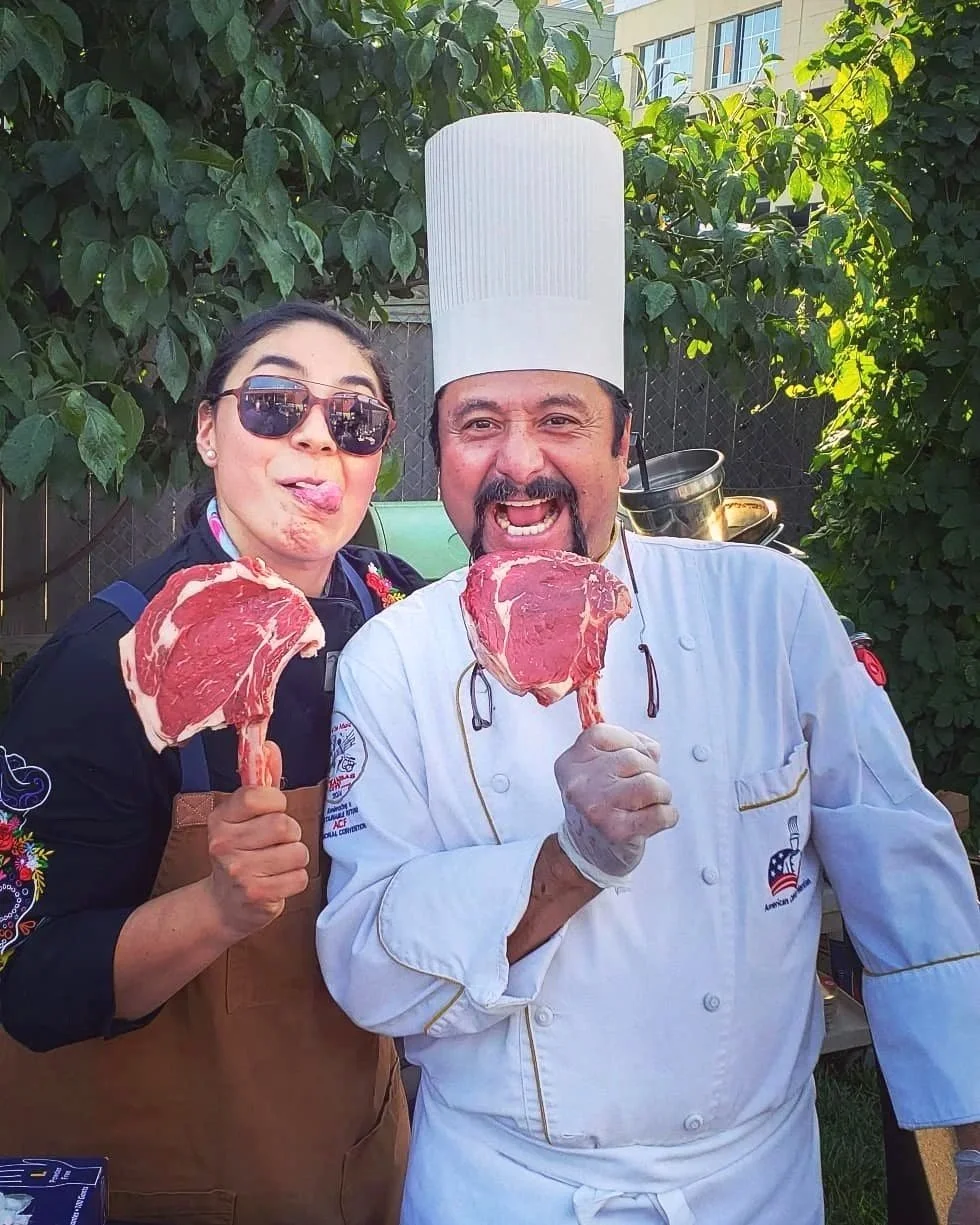 Two people, a woman and a man dressed as a chef, holding large raw steaks on skewers and smiling. The woman is wearing sunglasses and has her tongue out, while the chef is wearing a tall white hat and white chef coat, both outdoors with greenery in the background.