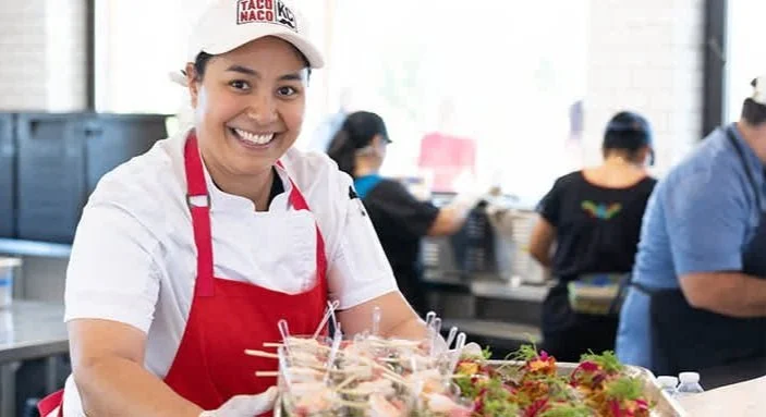 Chef Fernanda Reyes in a white cap and red apron smiling at a food counter at Taco Naco restaurant with other workers visible in the background.