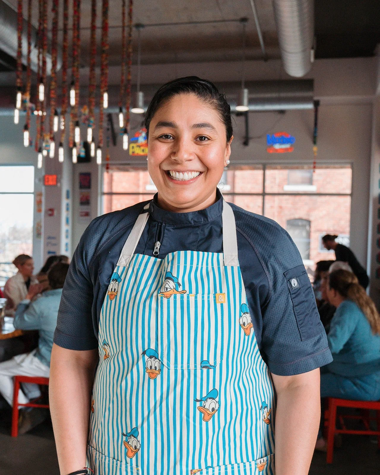 A smiling Chef Fernanda Reyes wearing a blue striped apron with Donald Duck's face at a restaurant or cafe.