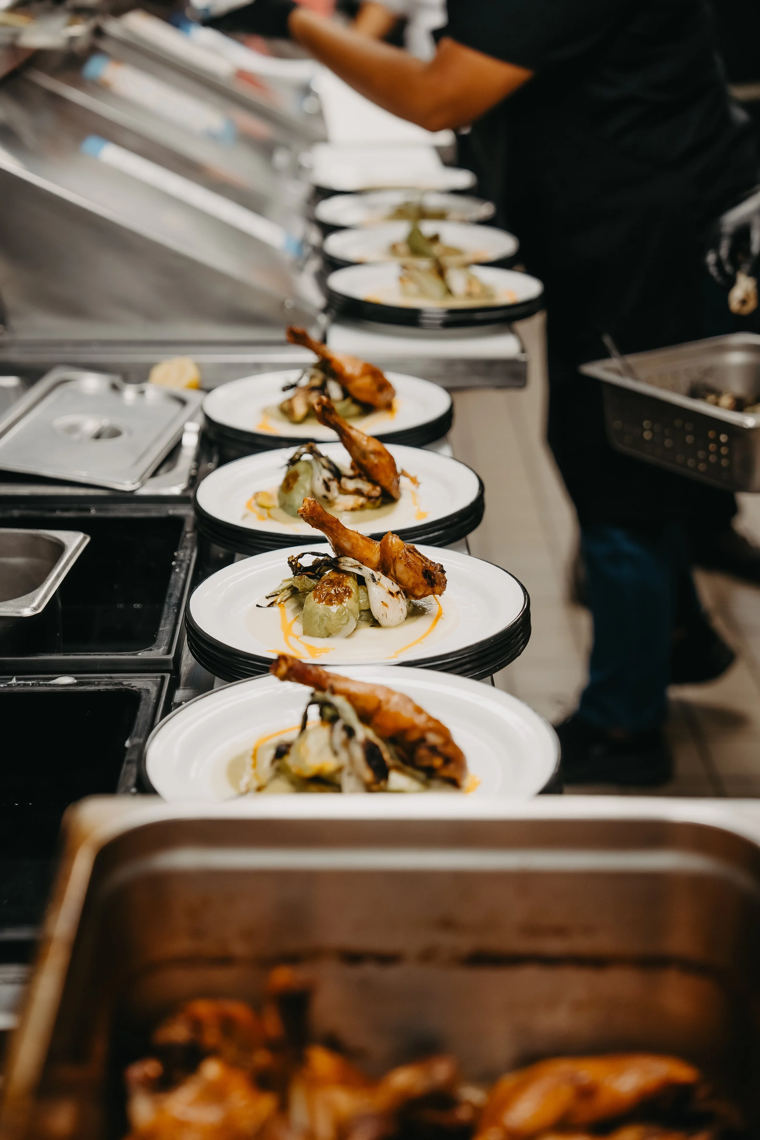 Plates with roasted chicken drumsticks and vegetables lined up on a kitchen counter, with a person preparing food in the background.