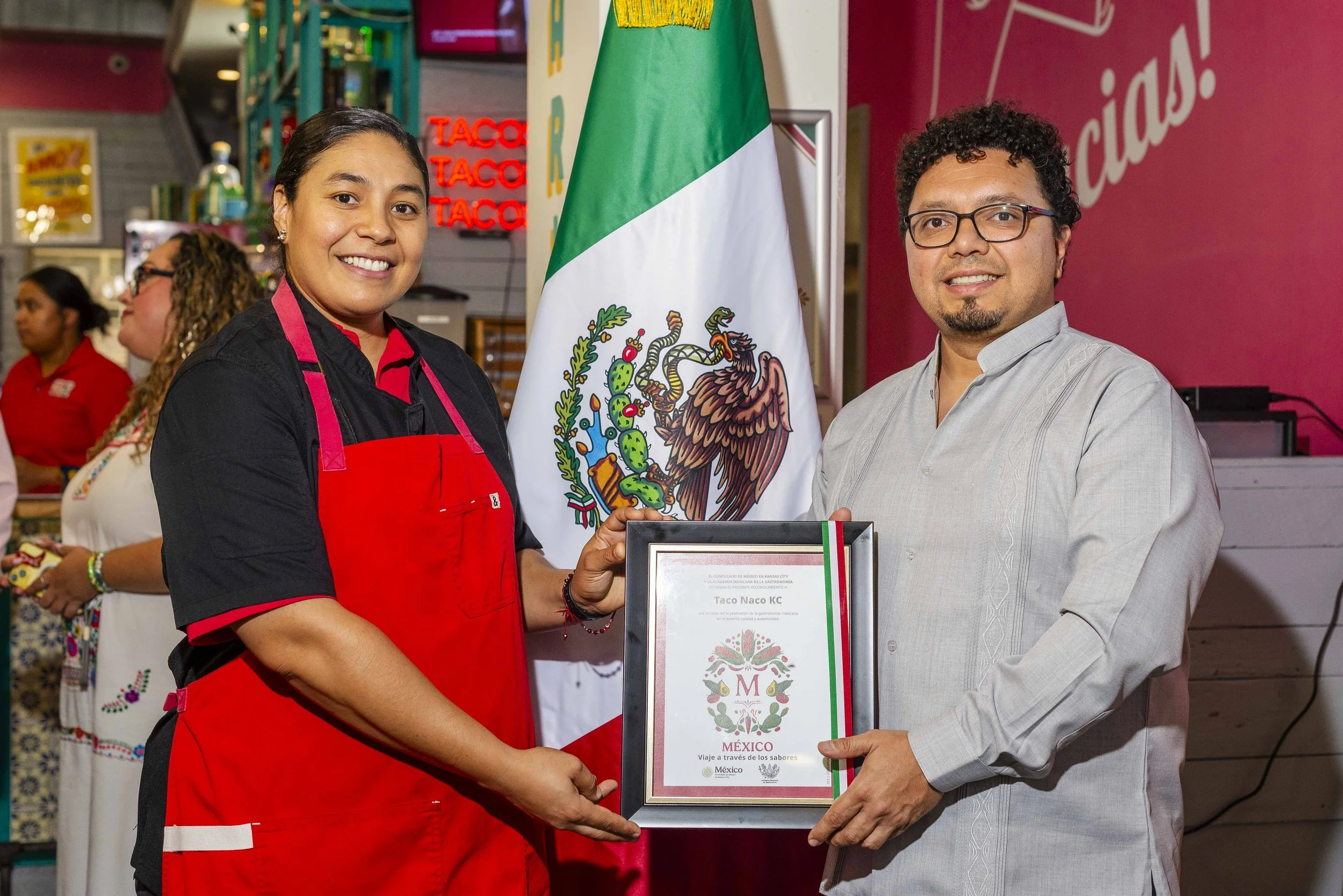 A woman in a black and red uniform is accepting an award from a man in a gray shirt. They are standing in front of the Mexican flag at an event, with other people in the background. The woman is smiling and holding a plaque, while the man is also smiling.
