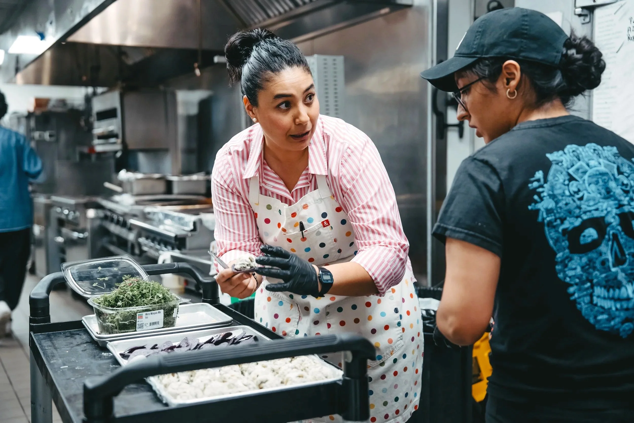 A woman wearing an apron with colorful polka dots is having a conversation with another person in a kitchen. The woman is holding a small object, possibly a piece of food, and there are containers of greens and other ingredients on the counter in front of them.