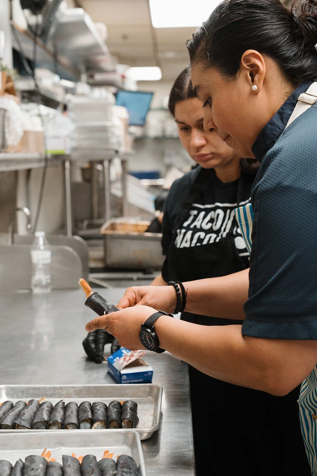 Two women preparing a chef-forward dish in a professional kitchen, with trays of sushi rolls and ingredients on the stainless steel counter.