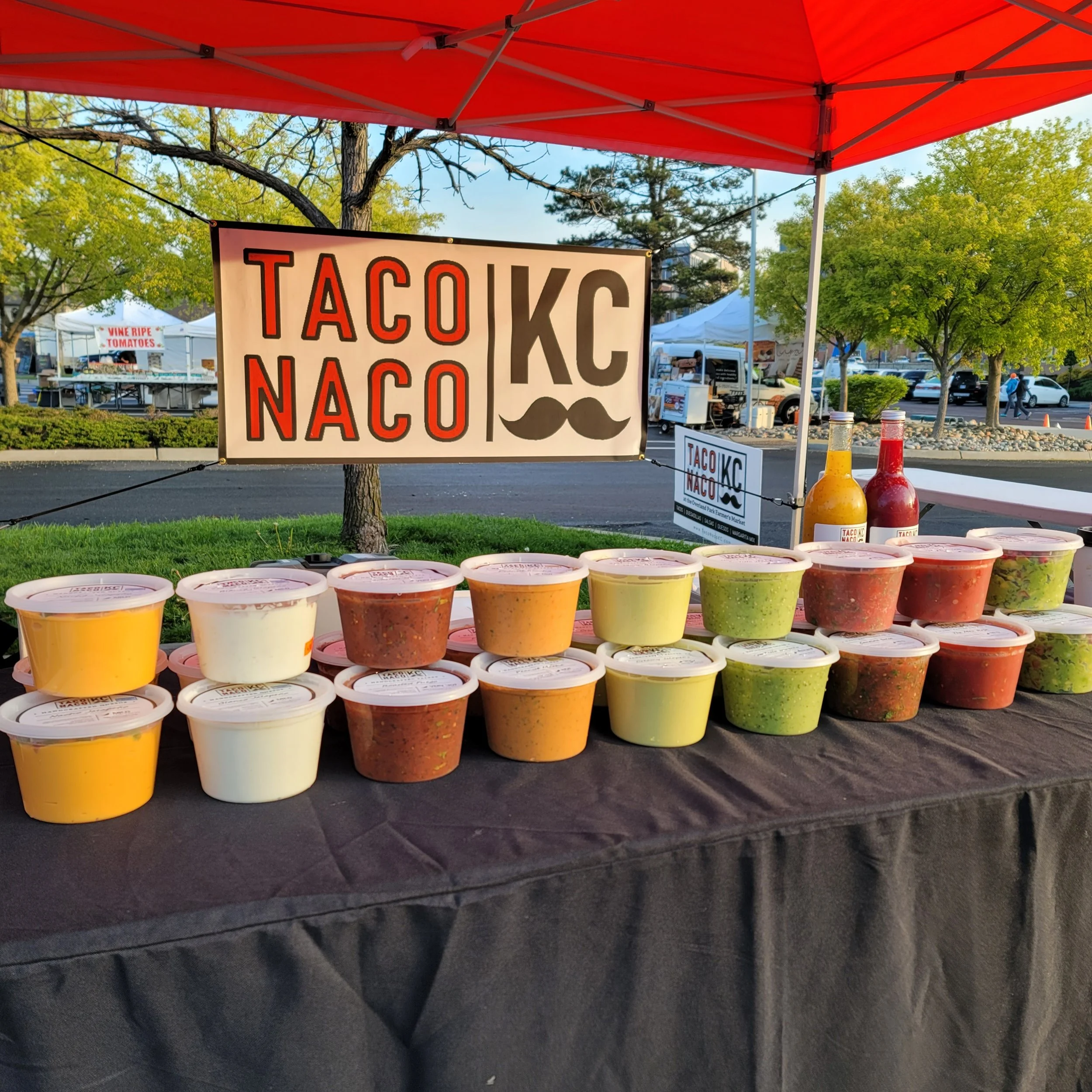 Taco Naco KC market stall displaying colorful containers of salsa, sauces, and guacamole under a red canopy