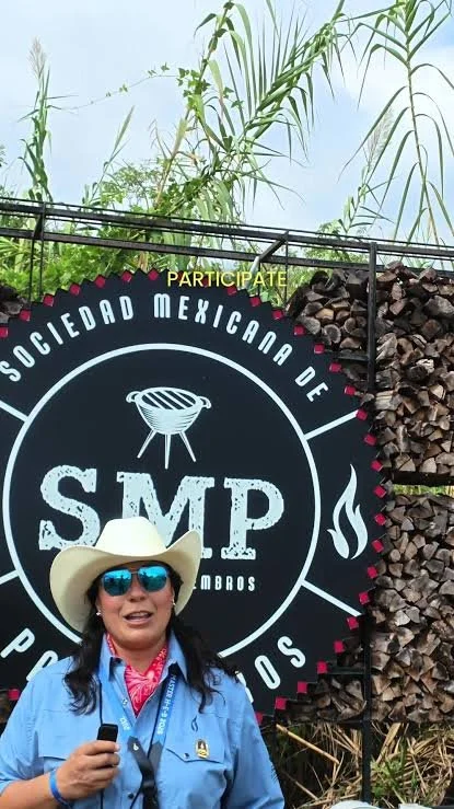 Woman wearing a cowboy hat and sunglasses, holding a smartphone. She is standing in front of a sign that reads 'Sociedad Mexicana de CMP' with a graphic of a grill and fire, and another word partially visible, likely 'Participate'.