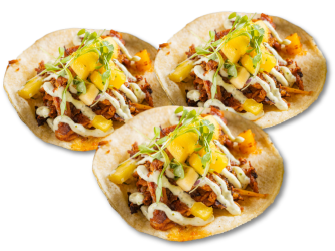 Three tacos filled with shredded meat, diced yellow peppers, shredded cheese, microgreens, with a drizzle of white sauce, on a black background.